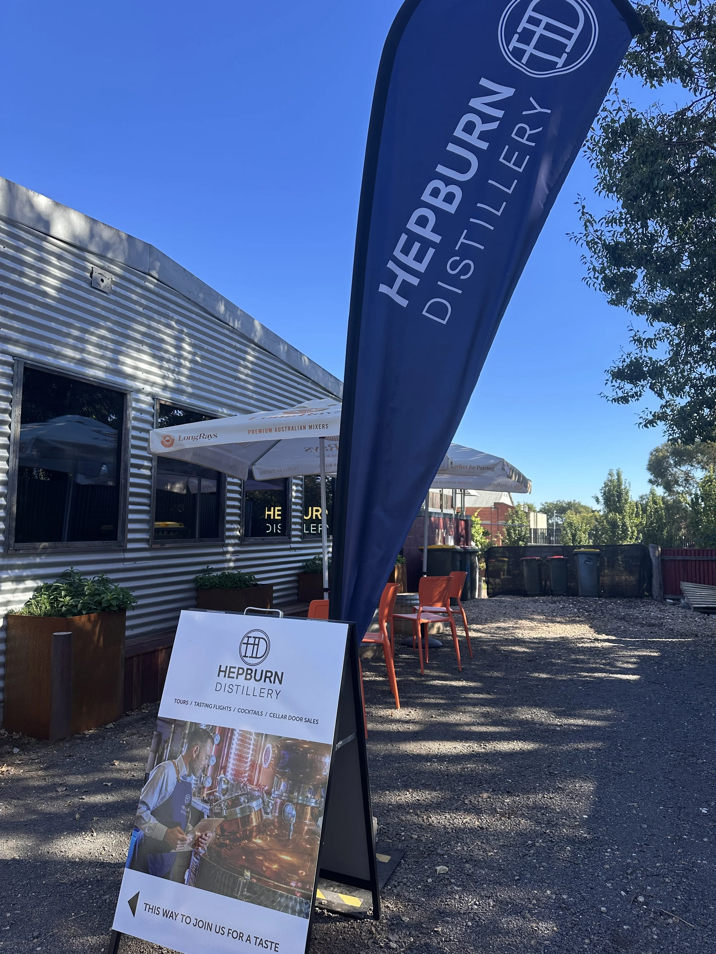 Outdoor signage and banner for Hepburn Distillery with a photo of a bartender in a distillery, set against a building with metal siding, trees, and seating area.