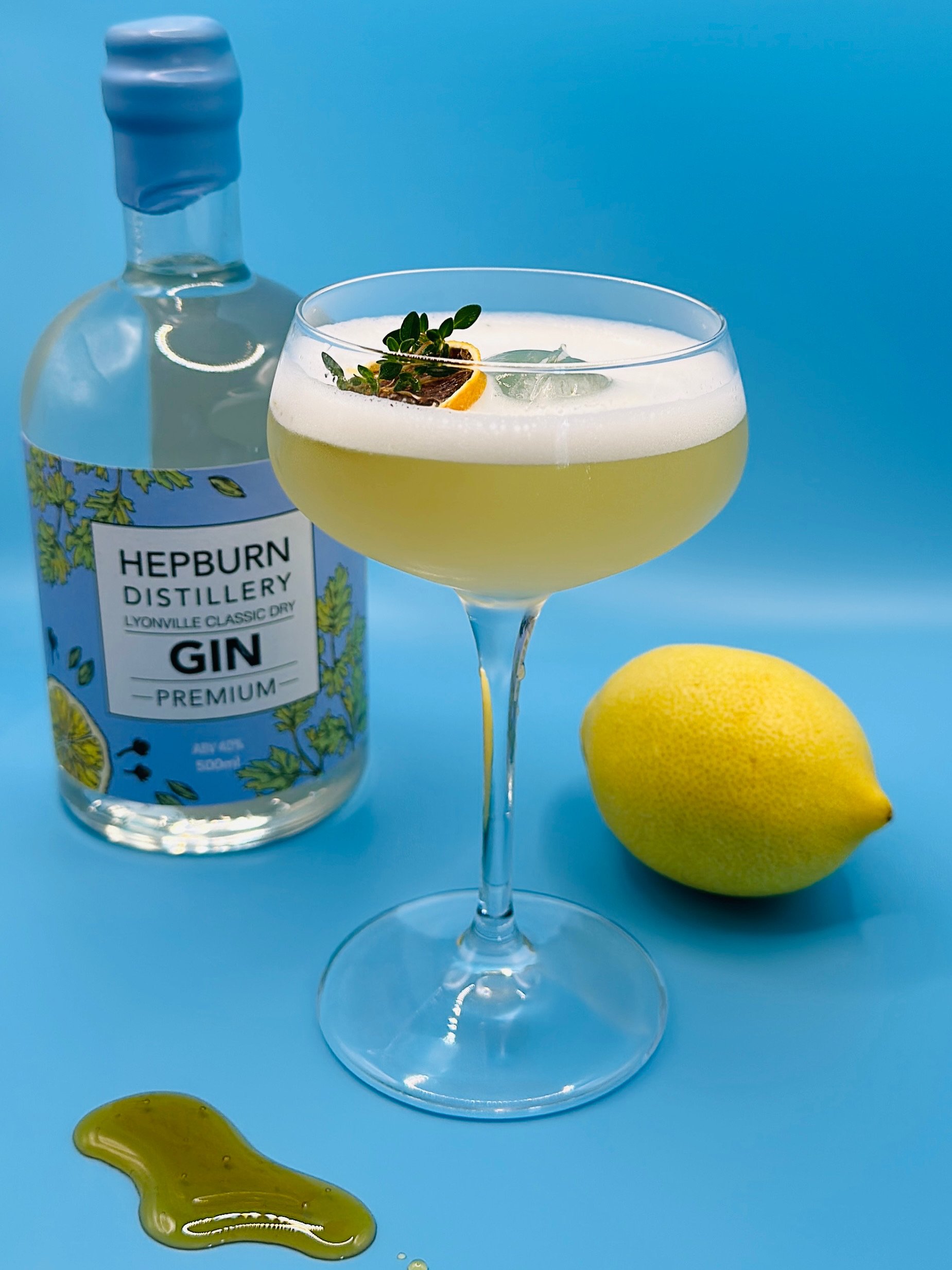 A lemon gin cocktail garnished with lemon zest and herbs in a coupe glass, with a bottle of Hepburn distillery gin, a lemon, and some honey on a blue background.