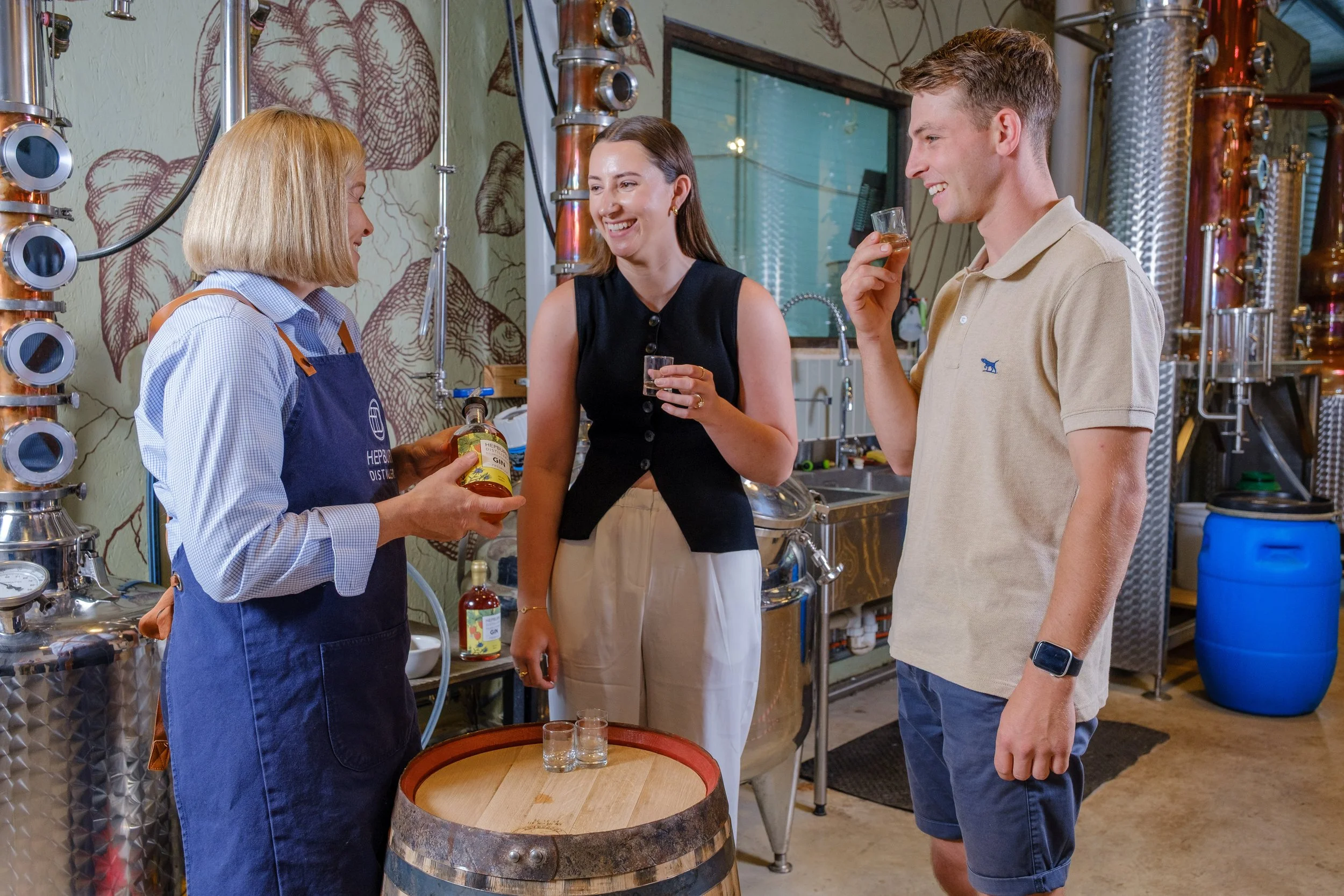Two women and one man tasting gin in a distillery, with a woman in an apron talking to the couple