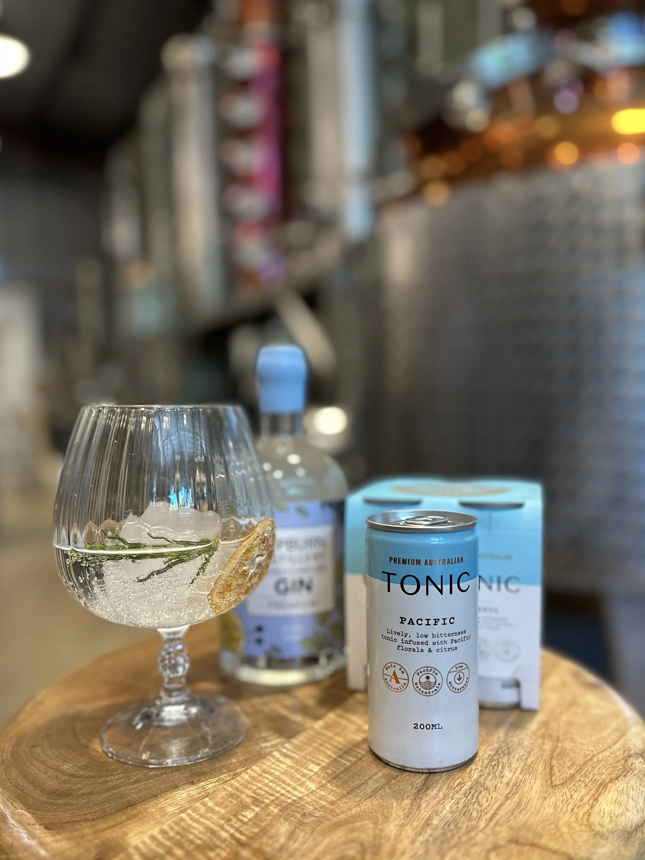 A glass of tonic water with ice and a slice of lemon, a can of Pacific tonic water, a box of Pacific tonic water, and a water bottle on a wooden table in a bar or kitchen setting.