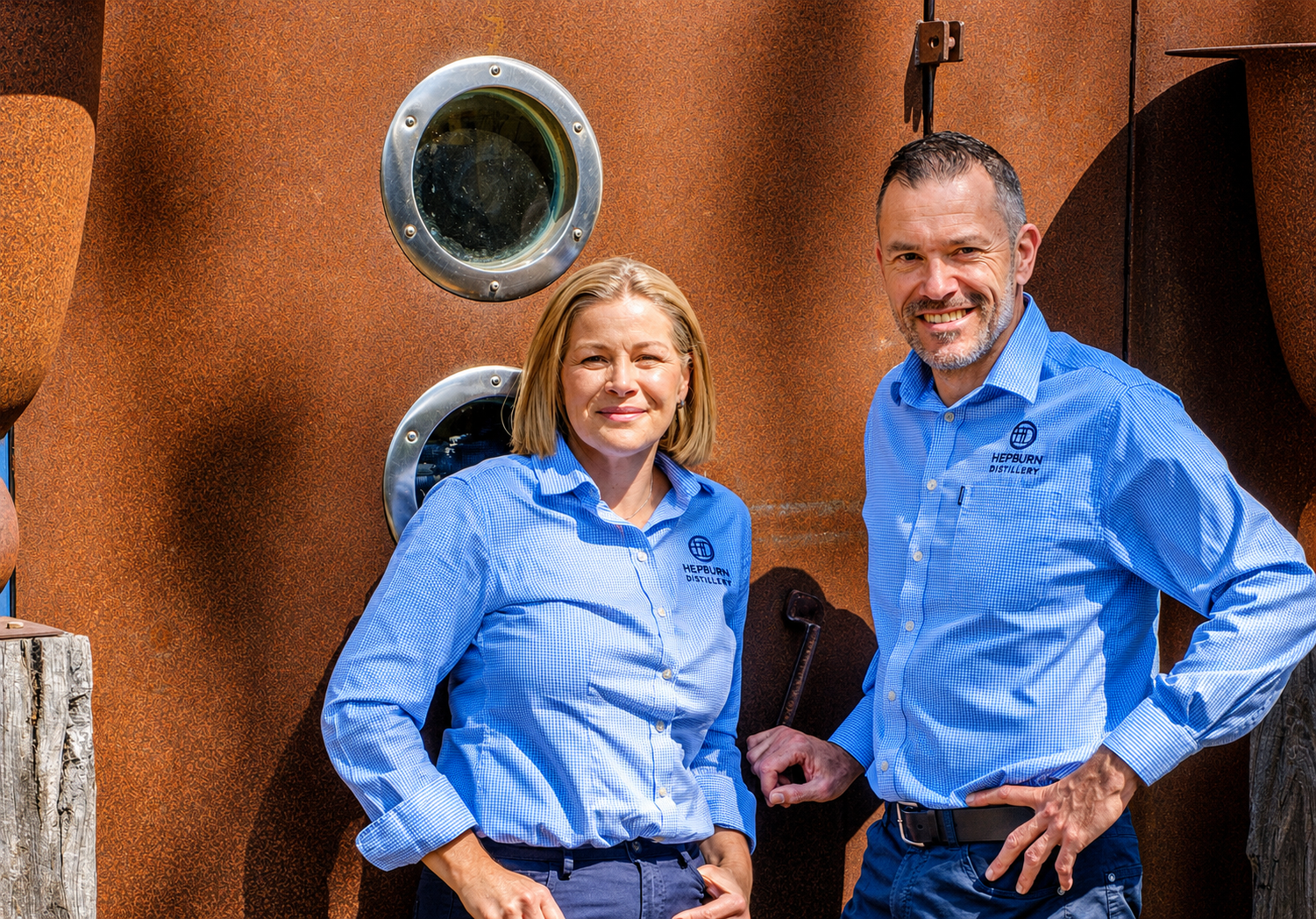 Two people, a woman and a man, standing outdoors in front of a rusty metal structure with two round windows. They are both wearing blue shirts embroidered with 'Hepburn Distillery.' The woman has blonde hair and is smiling slightly, with her hands in