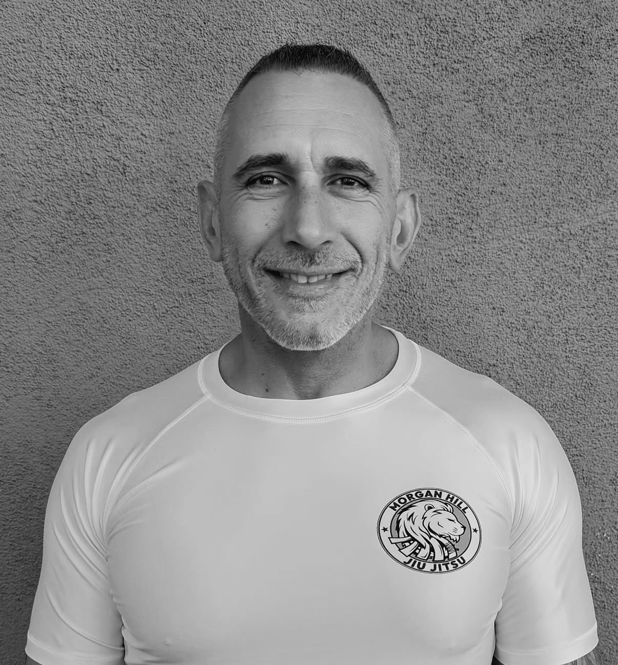 A man smiling in front of a wall, wearing a white T-shirt with a logo that reads "Morgan Hill Jiu Jitsu" and features a lion's head.