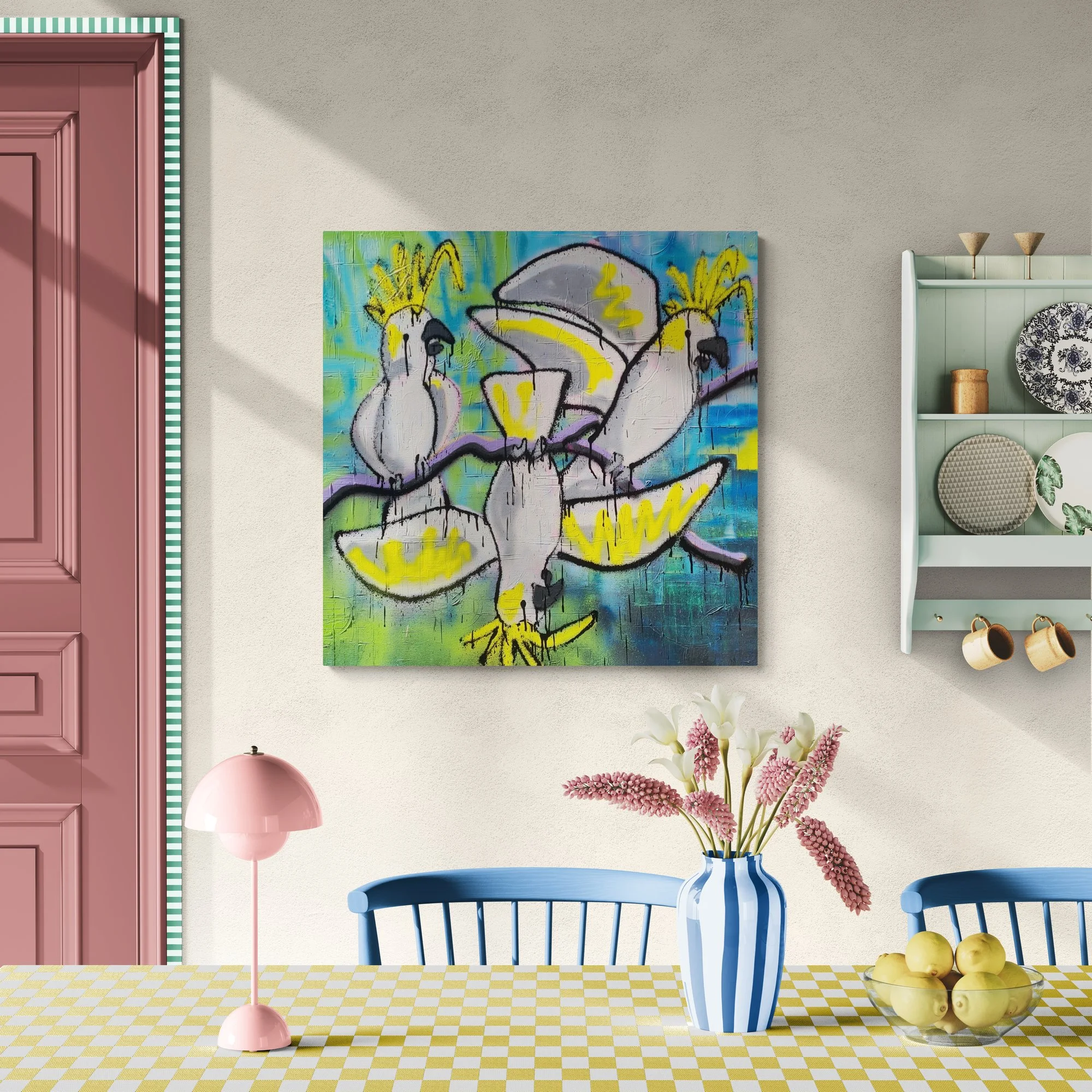 Colorful abstract painting of cockatoos on the wall above a dining table with a blue and white striped vase of flowers and a bowl of fruit, pink lamp, pink door, and shelves with dishes and cups.