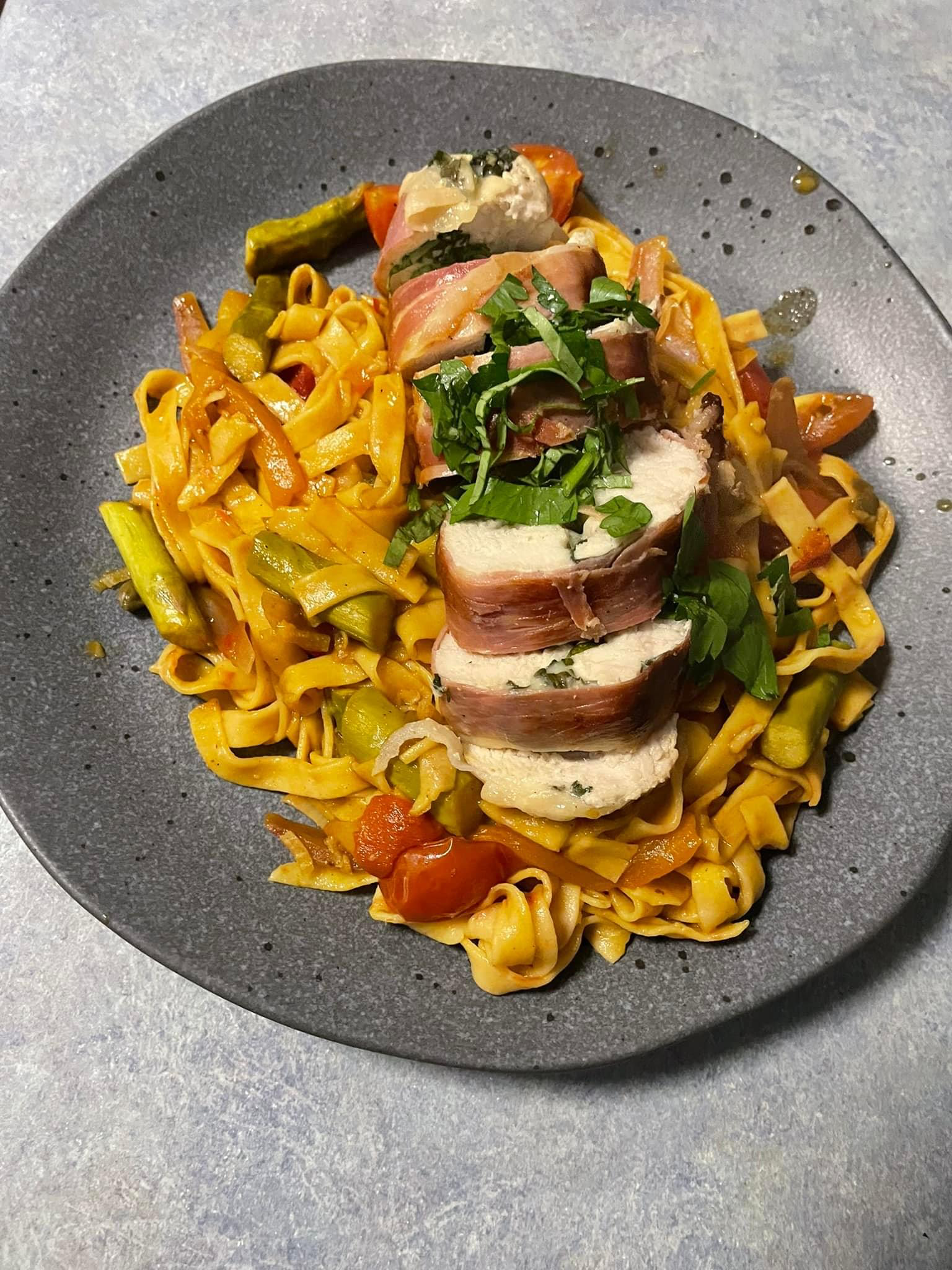 Plate of cooked pasta with vegetables topped with stuffed chicken breast wrapped in bacon, garnished with chopped parsley.