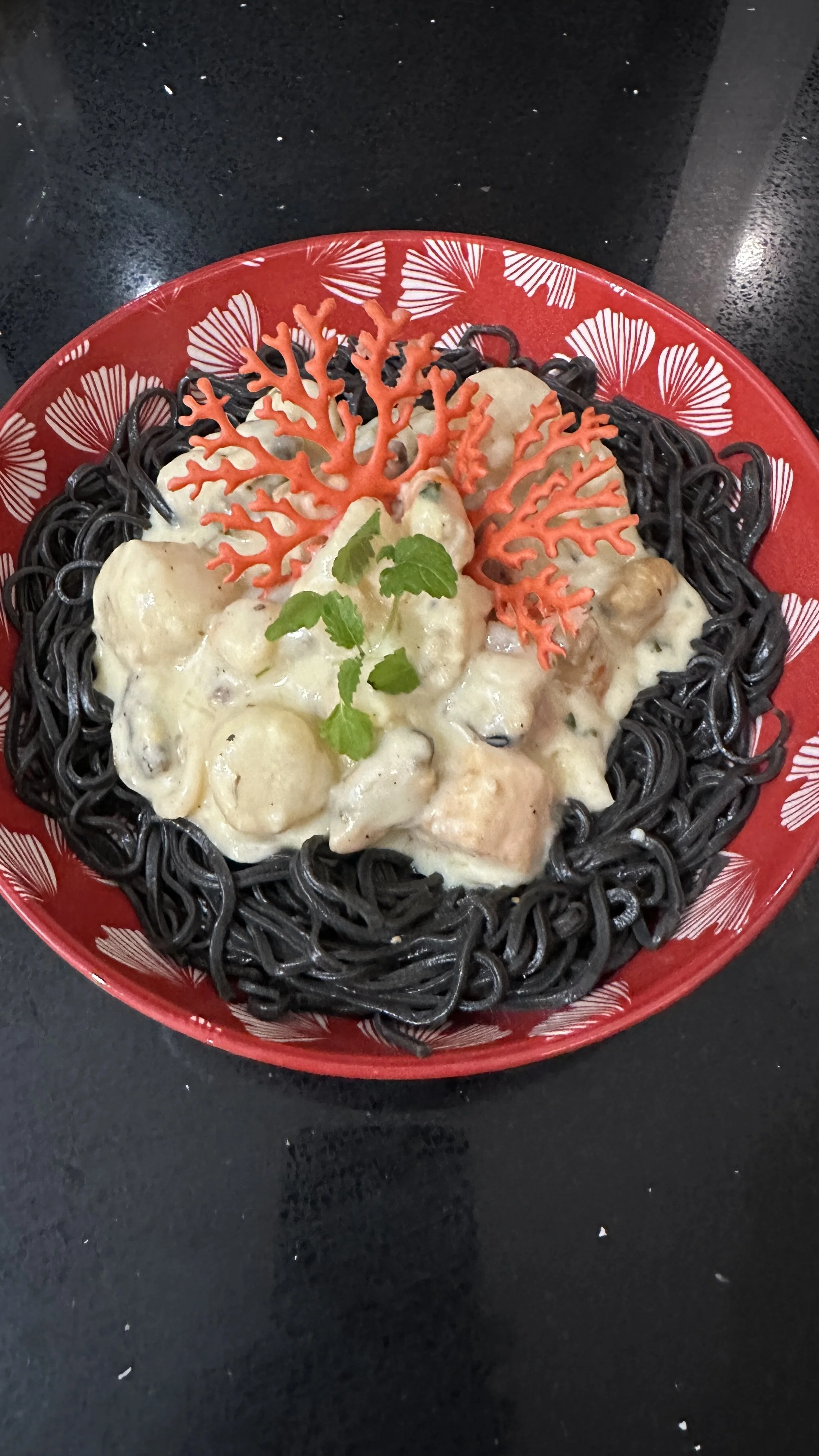 A bowl of black squid ink pasta topped with a creamy seafood and vegetable mixture, garnished with red coral-shaped decoration and a sprig of green herbs