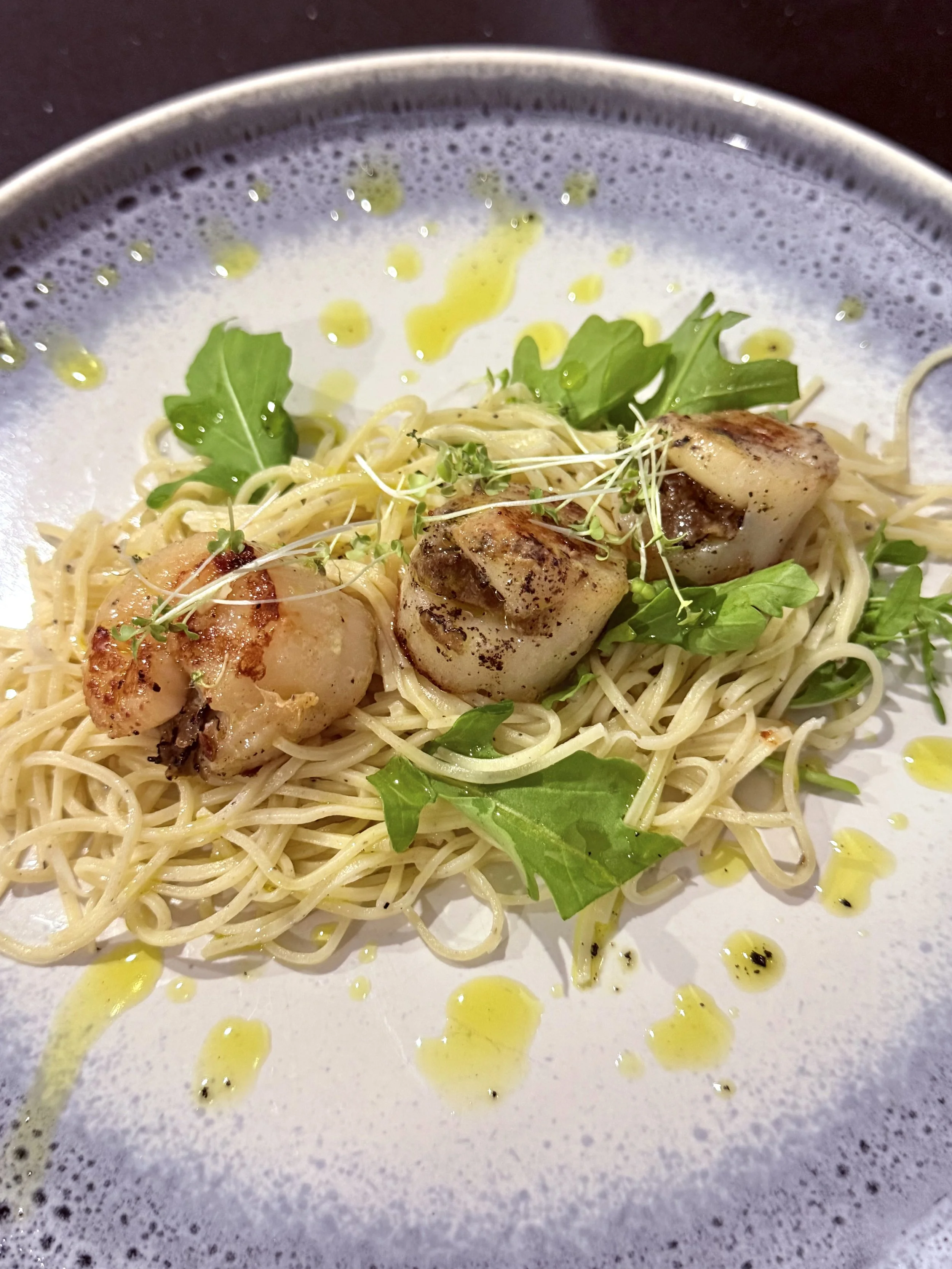 A plate of pasta with grilled scallops, garnished with microgreens and arugula, drizzled with olive oil, served on a gray ceramic plate.