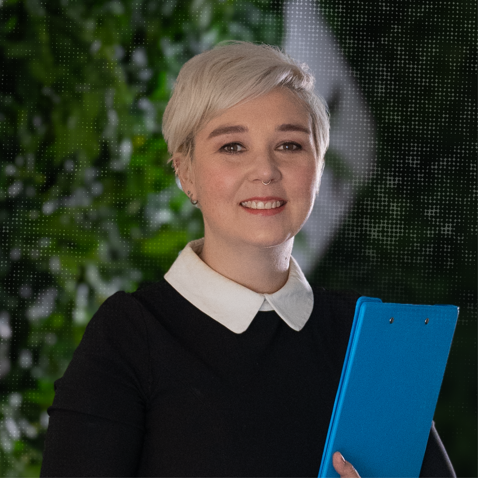 A woman with short blonde hair, wearing a black top with a white collar, holding a blue folder, smiling indoors with greenery in the background.