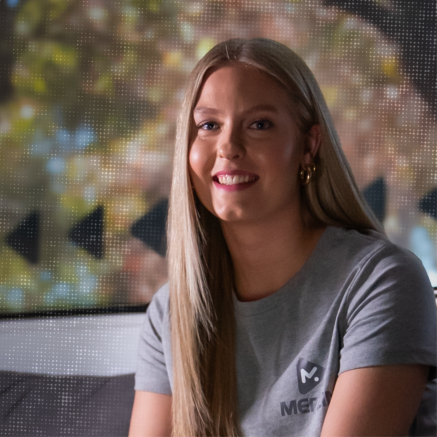 A young woman with long blonde hair smiling at the camera, wearing a gray t-shirt with a logo, in front of a festive, blurred background with Christmas lights and decorations.