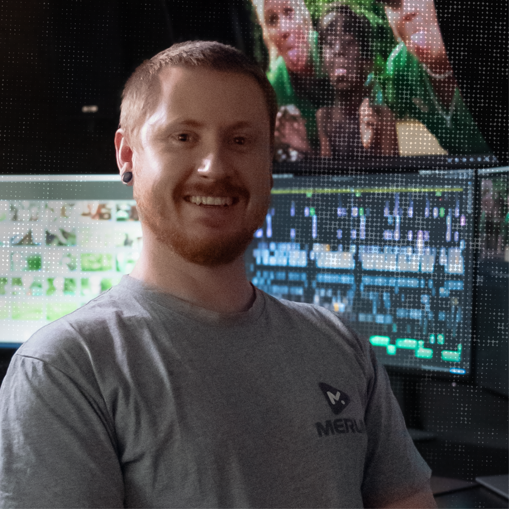 A smiling man with short red hair and tattoos on his arms standing in front of multiple computer monitors displaying graphs and images, in a dark room.