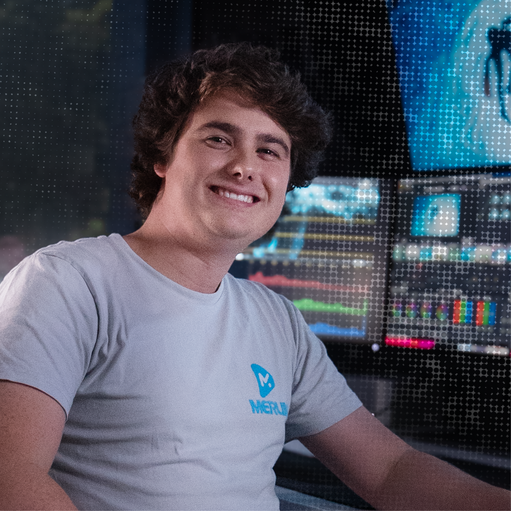 A smiling young man with curly hair in a gray T-shirt with a logo in front of multiple screens displaying colorful graphics, in a dark room.