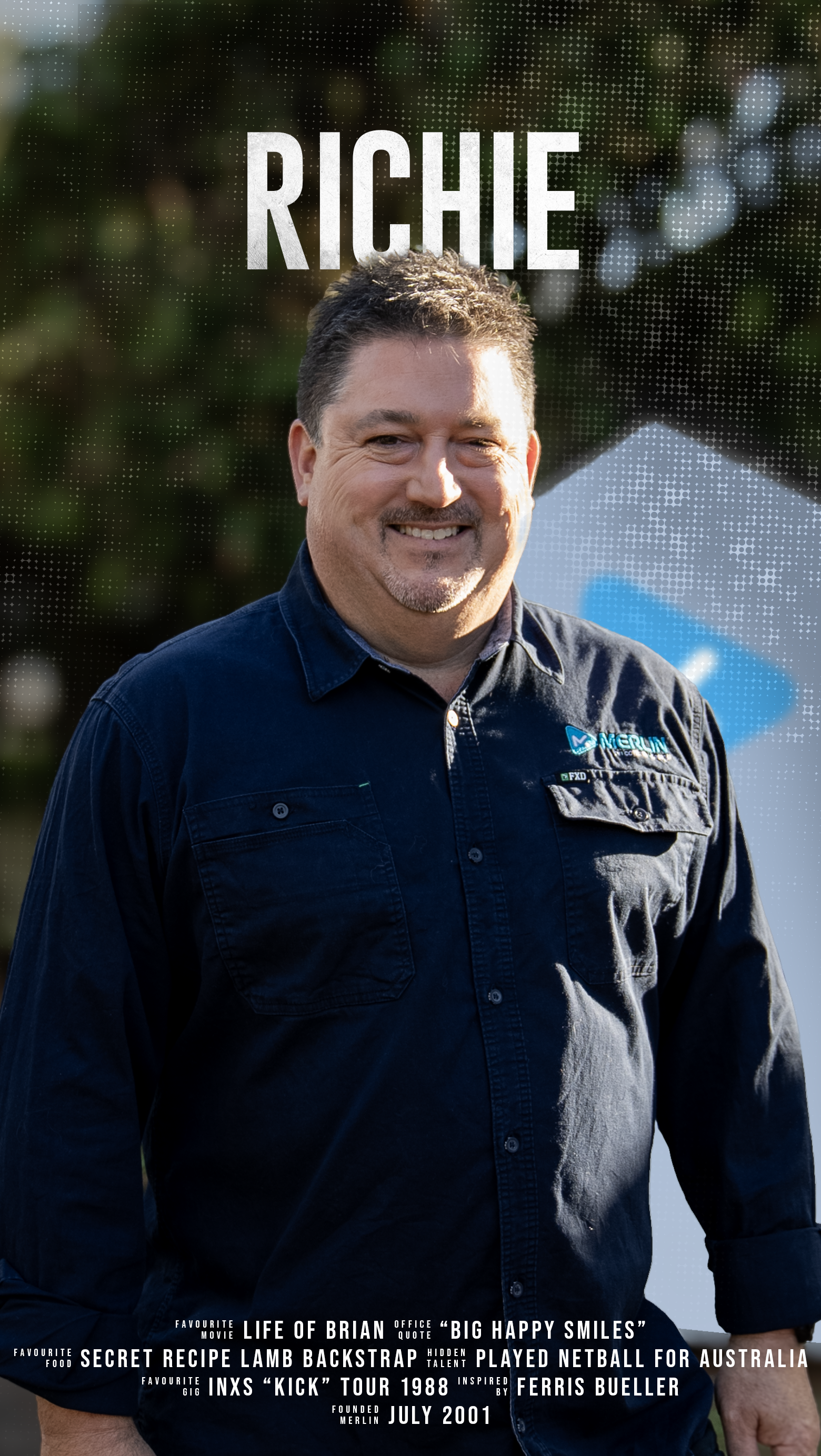 A man smiling outdoors with trees behind him, wearing a dark blue button-up shirt with a logo on the chest.