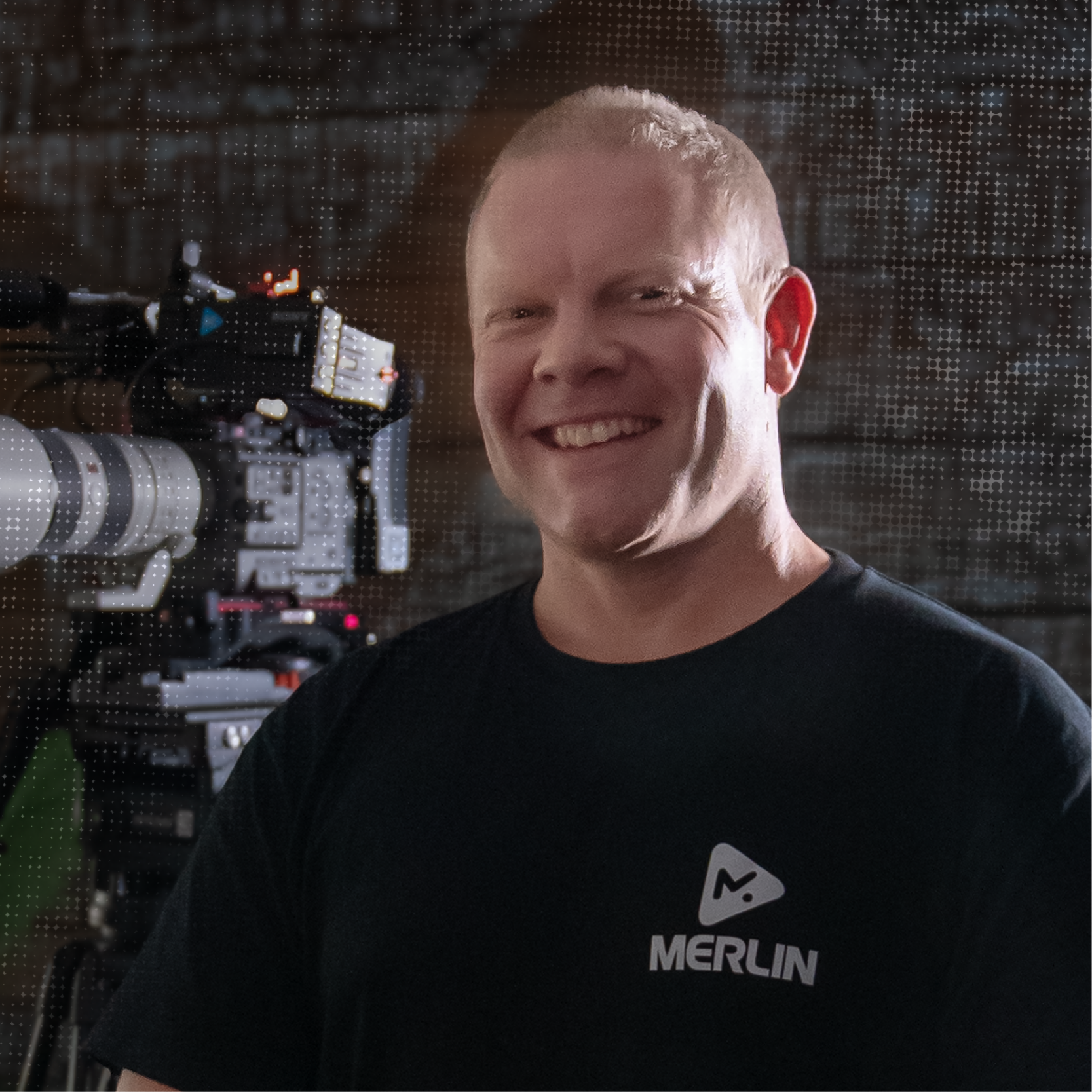 A man smiling at the camera in front of a professional camera setup, wearing a black t-shirt with 'Merlin' logo.