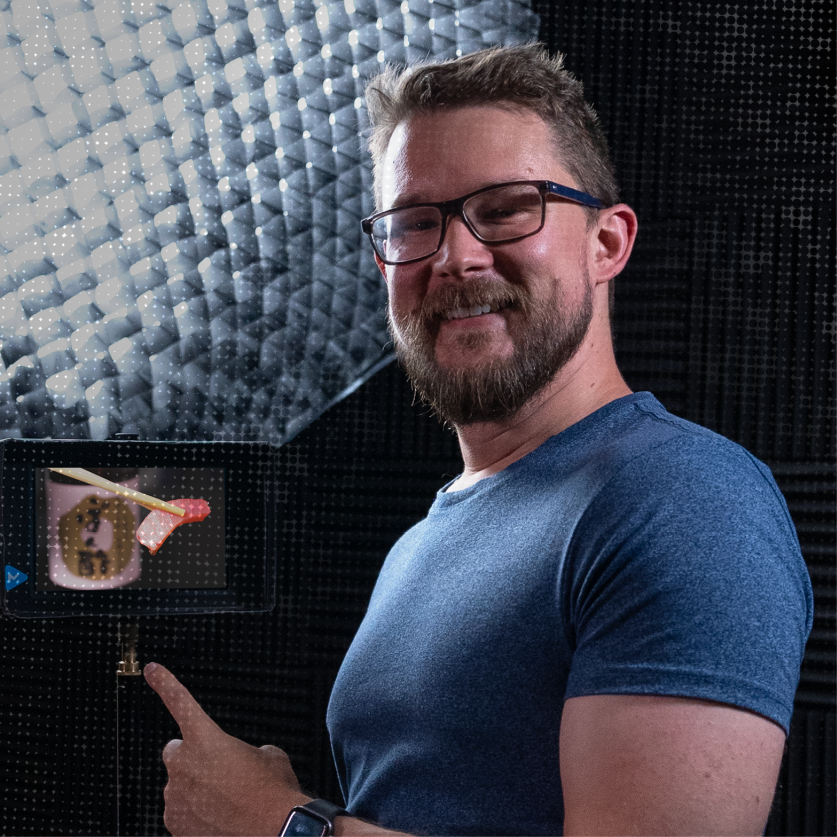 A man with glasses and a beard smiling and pointing at a small monitor showing a sushi roll and a piece of sushi with chopsticks.