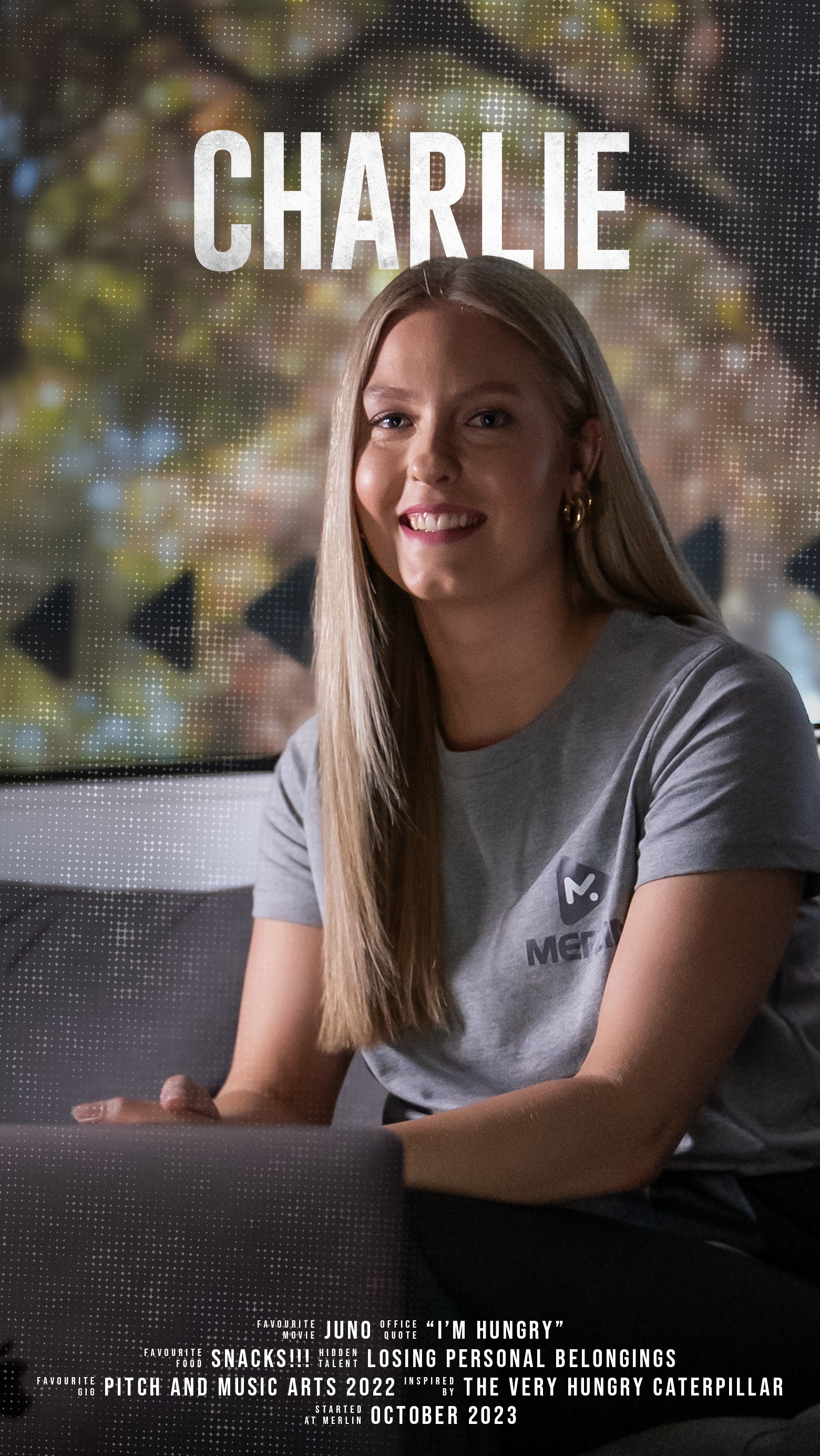 A young woman with long blonde hair, wearing a gray T-shirt, smiling while sitting indoors near a window with blurred holiday lights outside.