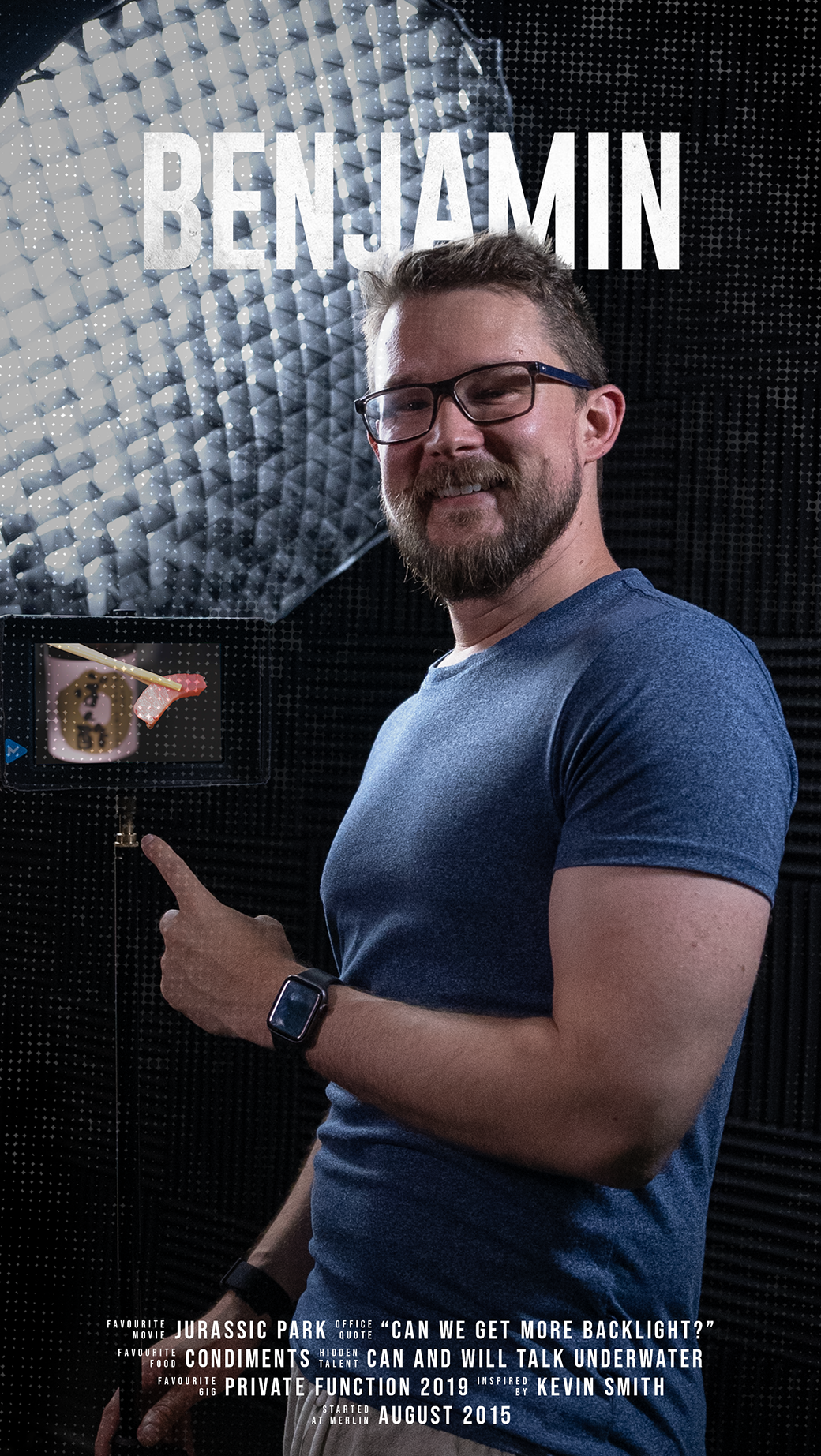 A man with glasses and a beard, wearing a blue t-shirt and a smartwatch, smiling and pointing at a small screen displaying food, in a recording studio or soundproof room, with the name 'Benjamin' displayed at the top.