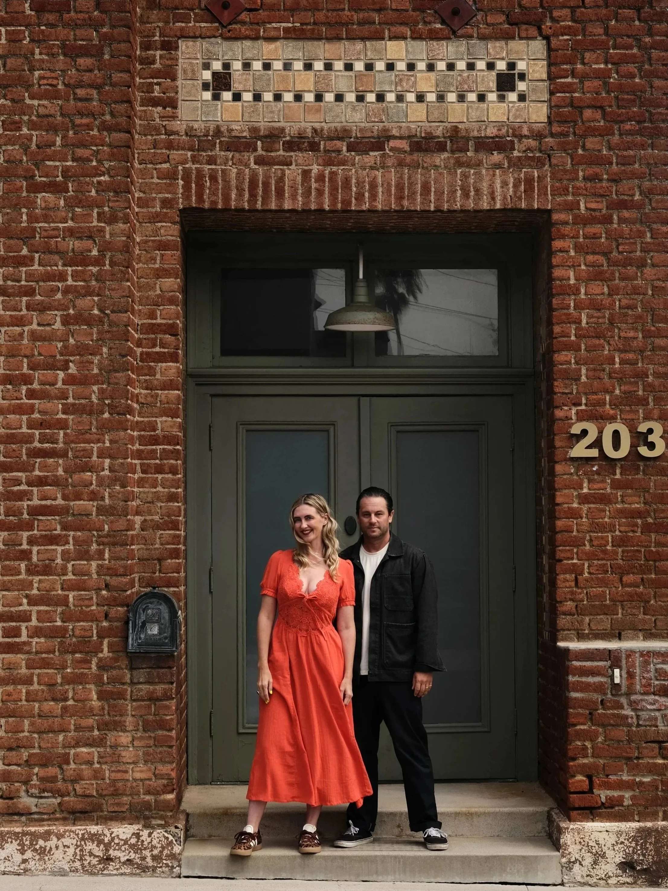 A couple standing in front of a brick building with a large door, their shadows cast on the ground, and the number 203 displayed on the wall to the right.