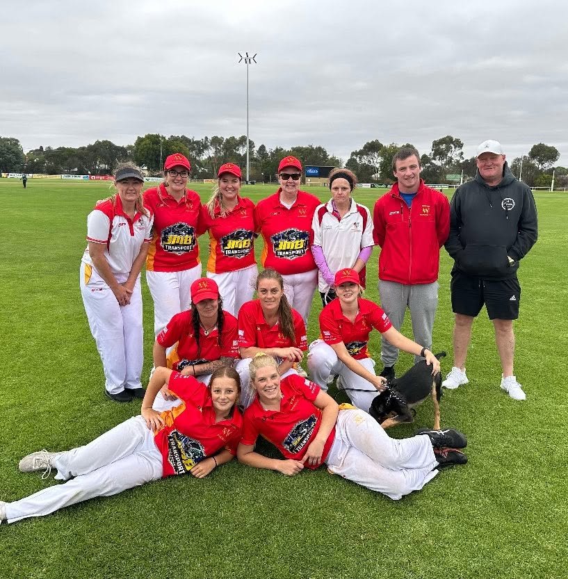 A group of women and men, some in red uniforms and others in casual attire, posing on a grassy sports field. Two women are lying on the ground in front, with a dog, and others are standing behind them. The overcast sky is visible in the background.