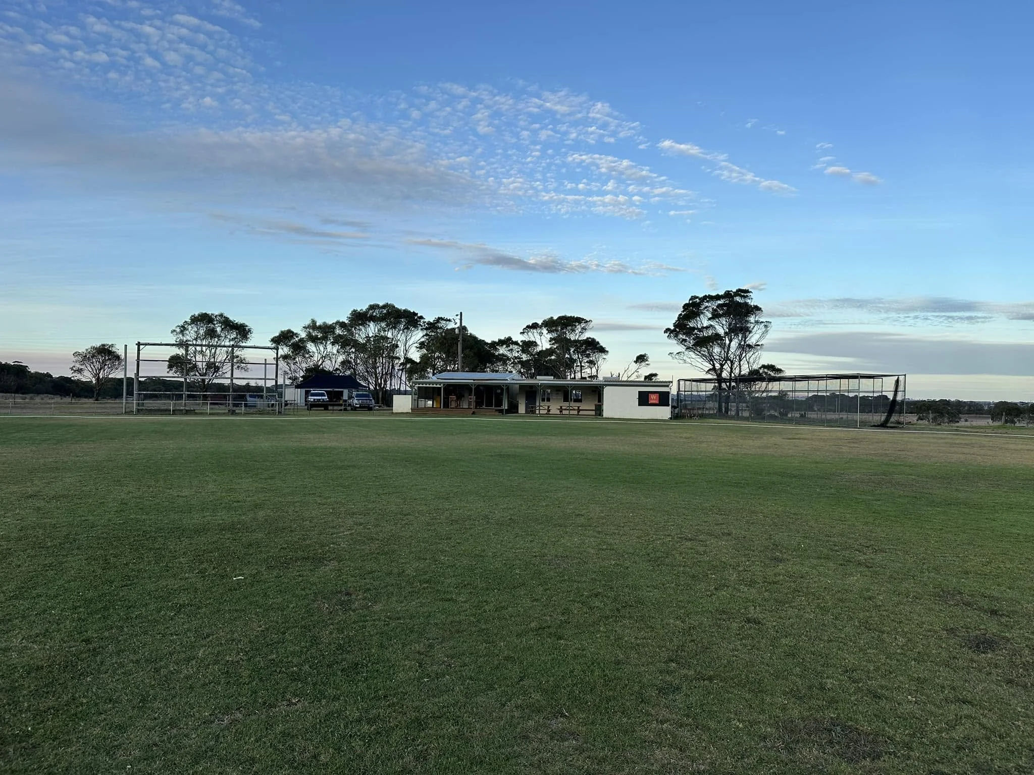 A sports field with green grass, trees in the background, and a small building or clubhouse under a partly cloudy sky.