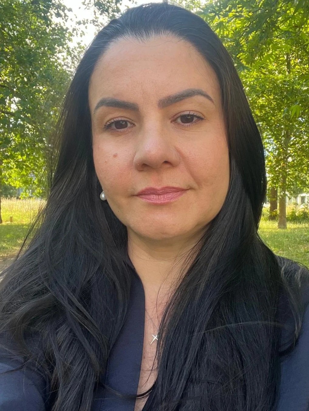 Close-up portrait of a woman with long black hair and pearl earrings, outdoors in a park with green trees and sunlight.