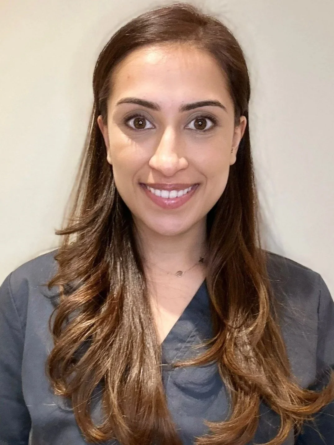 A young woman with long, wavy brown hair, wearing a dark top, smiling at the camera against a plain, light-colored background.