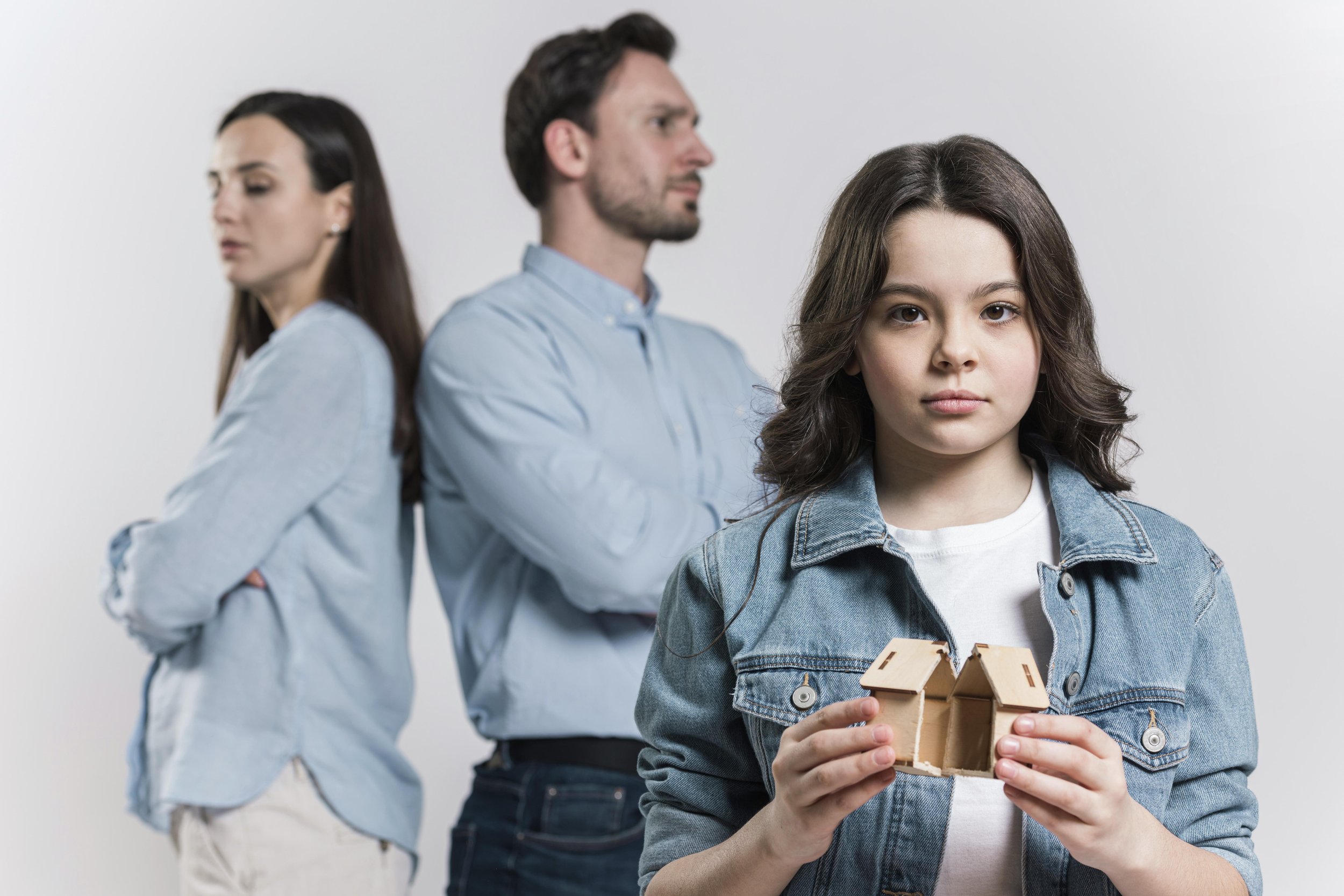 A young girl with dark wavy hair holding a small wooden house model, standing in front of a blurred background with a woman and a man, both with arms crossed, looking away.