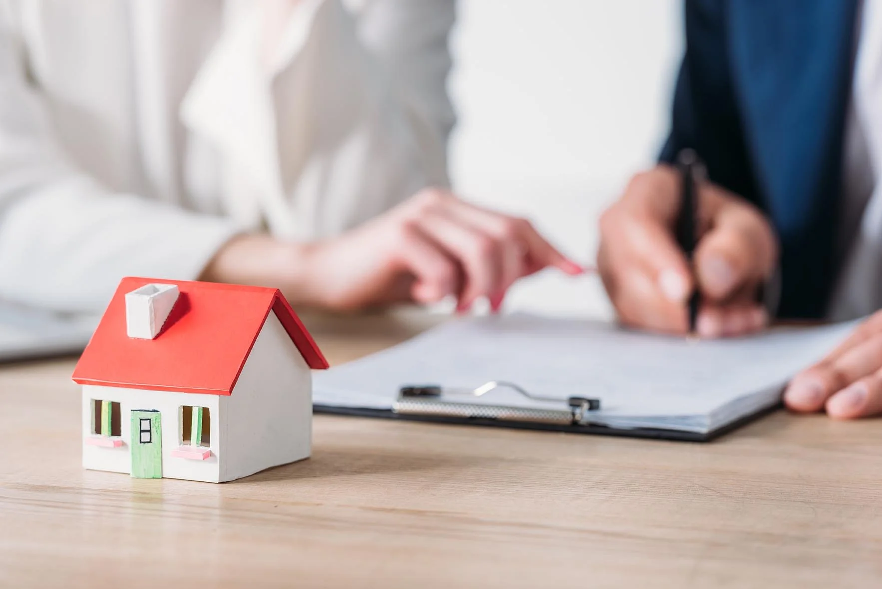 A miniature model of a house with a red roof on a wooden table in front of two people signing documents.