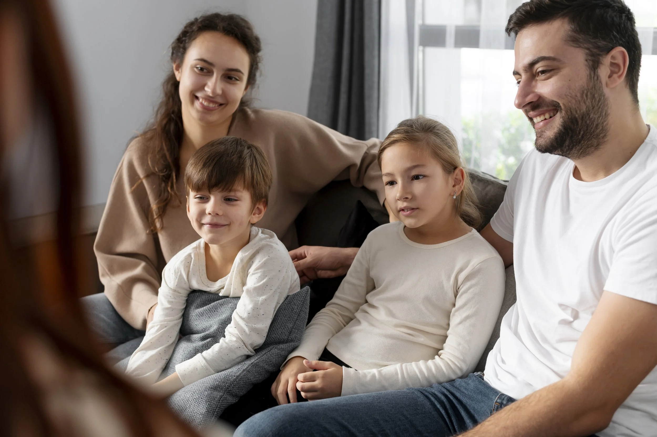 A family of four sitting on a couch and smiling during a video call in a living room with a window behind them.