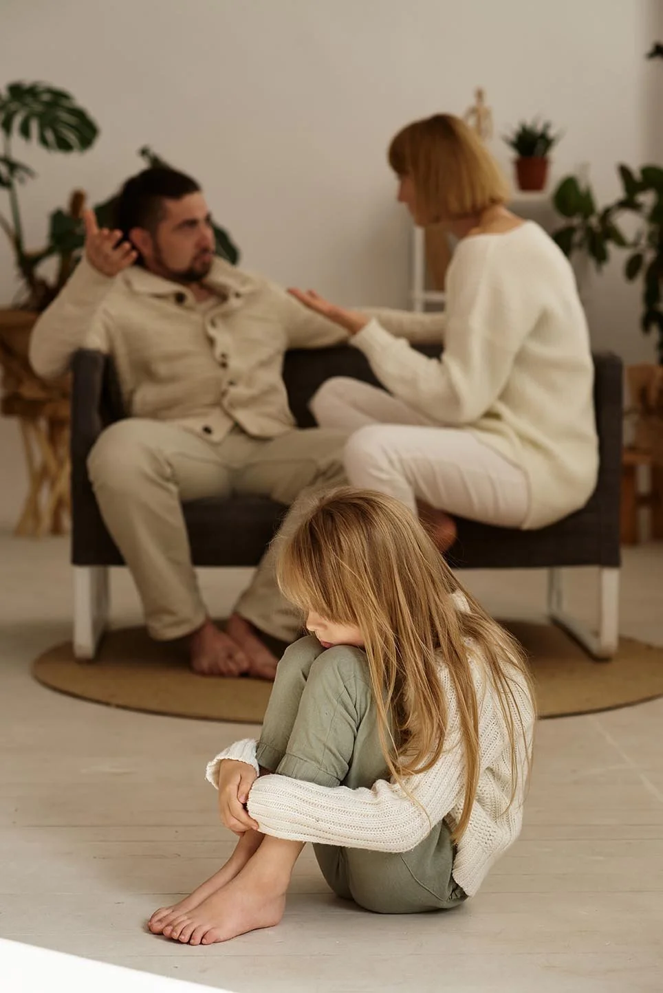 A woman and a man having a serious conversation on a gray sofa, a young girl sitting on the floor hugging her knees in the foreground.