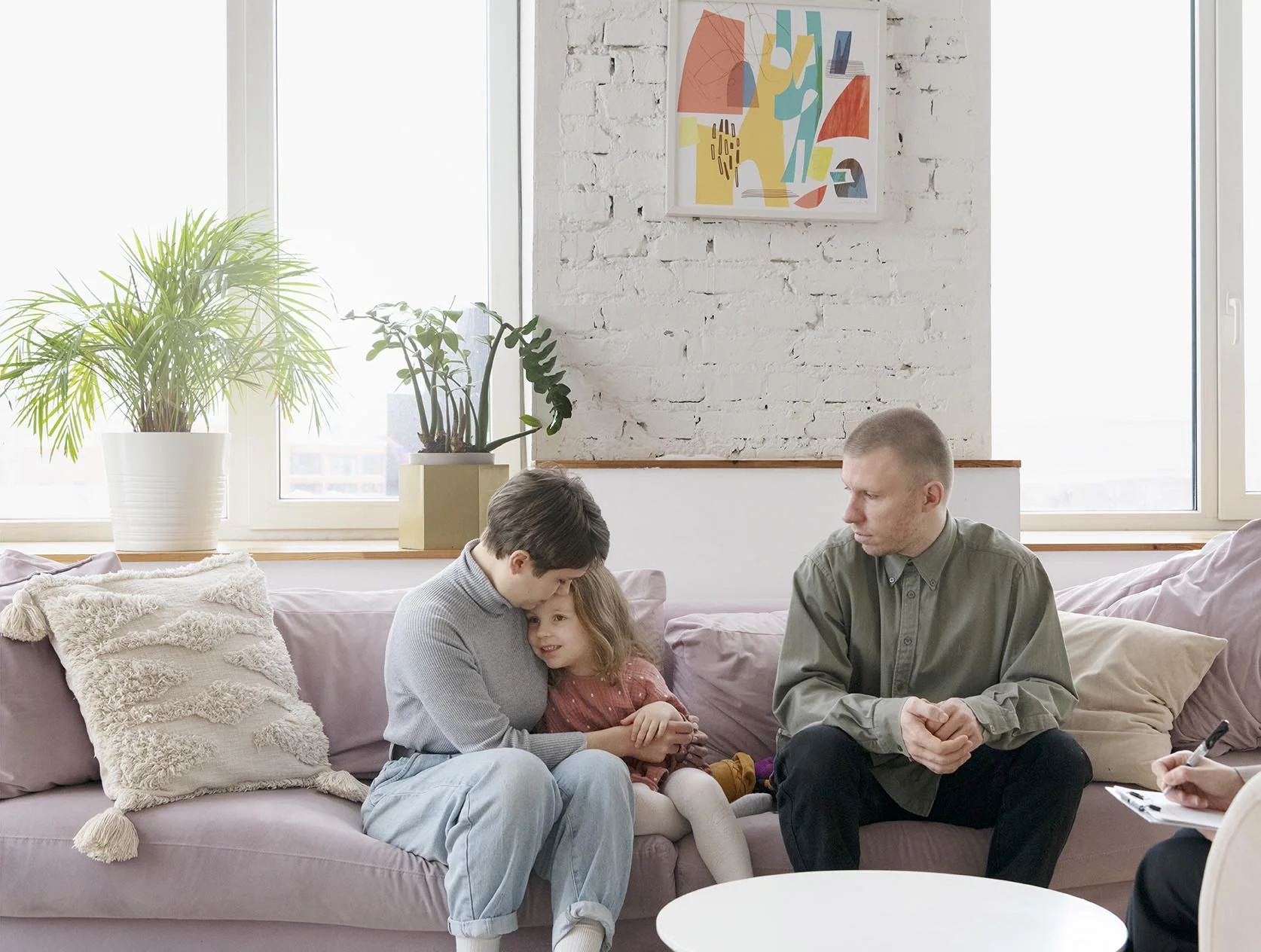 Family therapy session in a bright living room with white brick wall, colorful abstract art, large windows, and houseplants. A woman and a young girl sit close together on a pink sofa, with the woman comforting the girl. A man sits across, listening attentively. A therapist takes notes nearby.