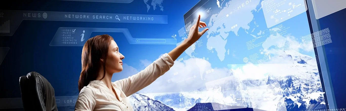 A woman with reddish-brown hair seated at a desk interacting with a large digital touchscreen display showing a world map and various digital interface elements.