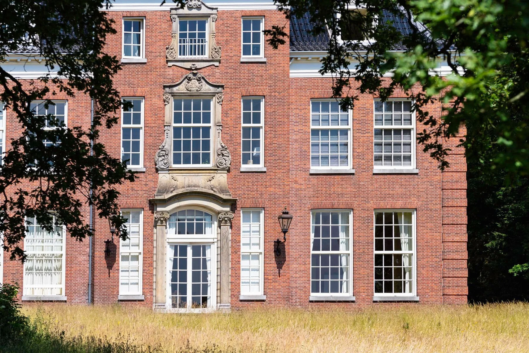 Een groot, rood-bruin bakstenen gebouw met witte kozijnen en een klassieke gevel, omgeven door groen gras en bomen.