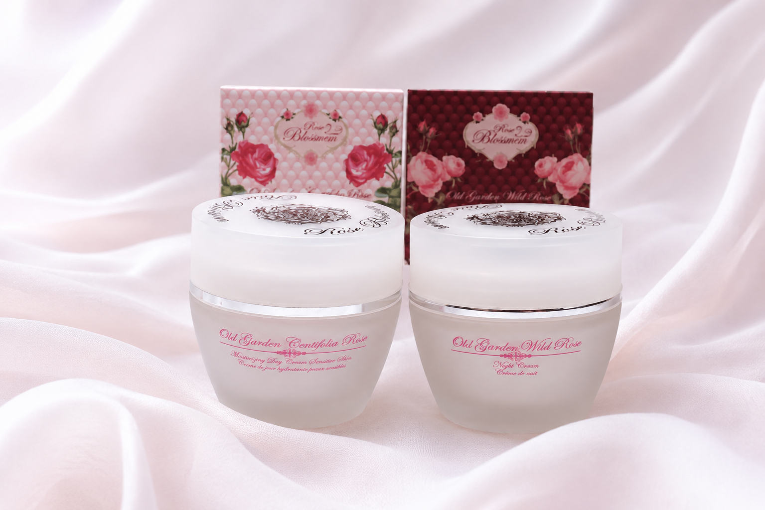 Two skincare jars labeled 'Old Garden Centifolia Rose' and 'Old Garden Wild Rose' with pink floral boxes in the background on a soft pink fabric surface.
