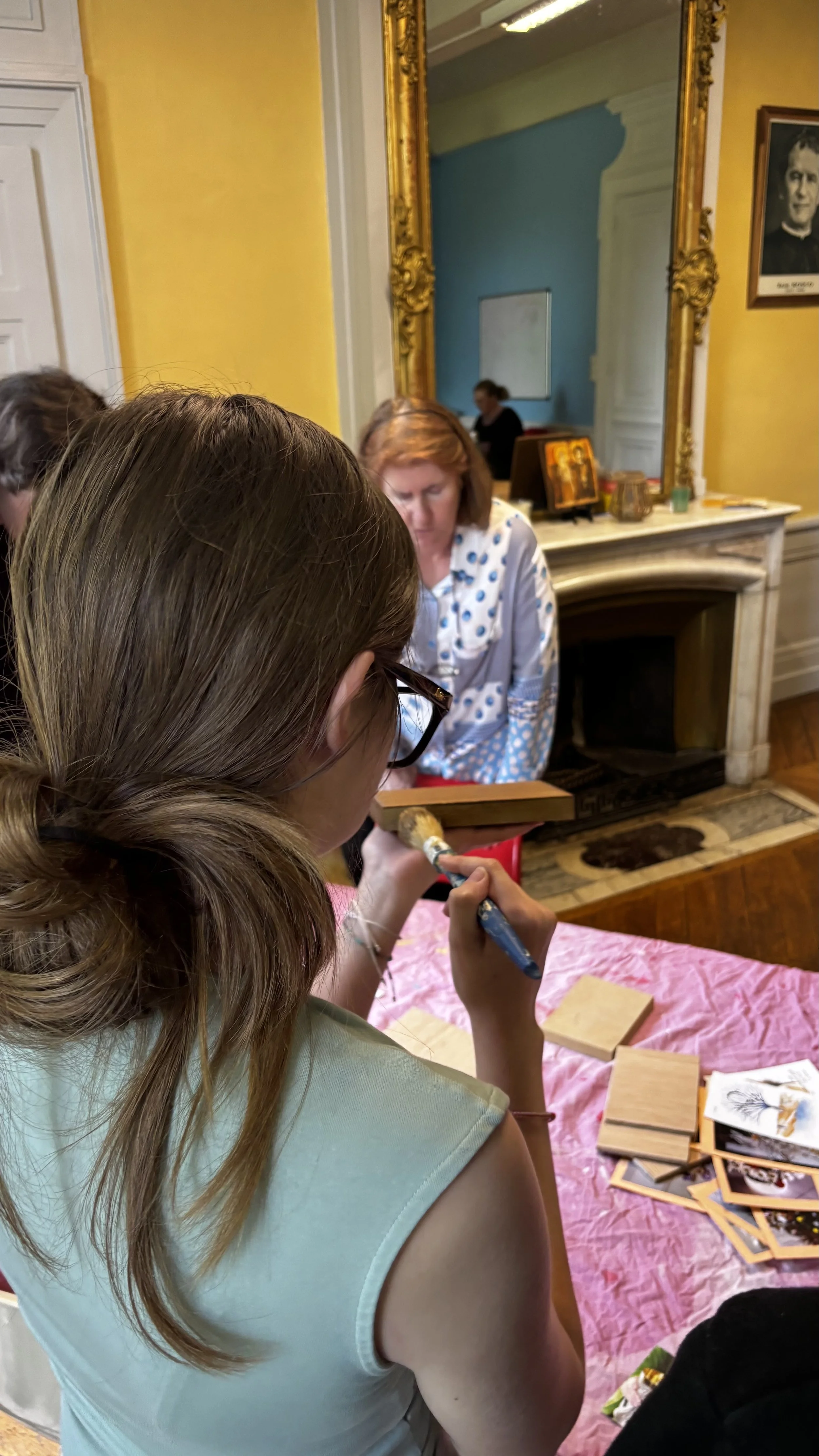 Une femme peignant une image dans une pièce ornée de couleurs vives, avec un grand miroir doré, un portrait en noir et blanc de personne dans le cadre et une cheminée blanche décorée.