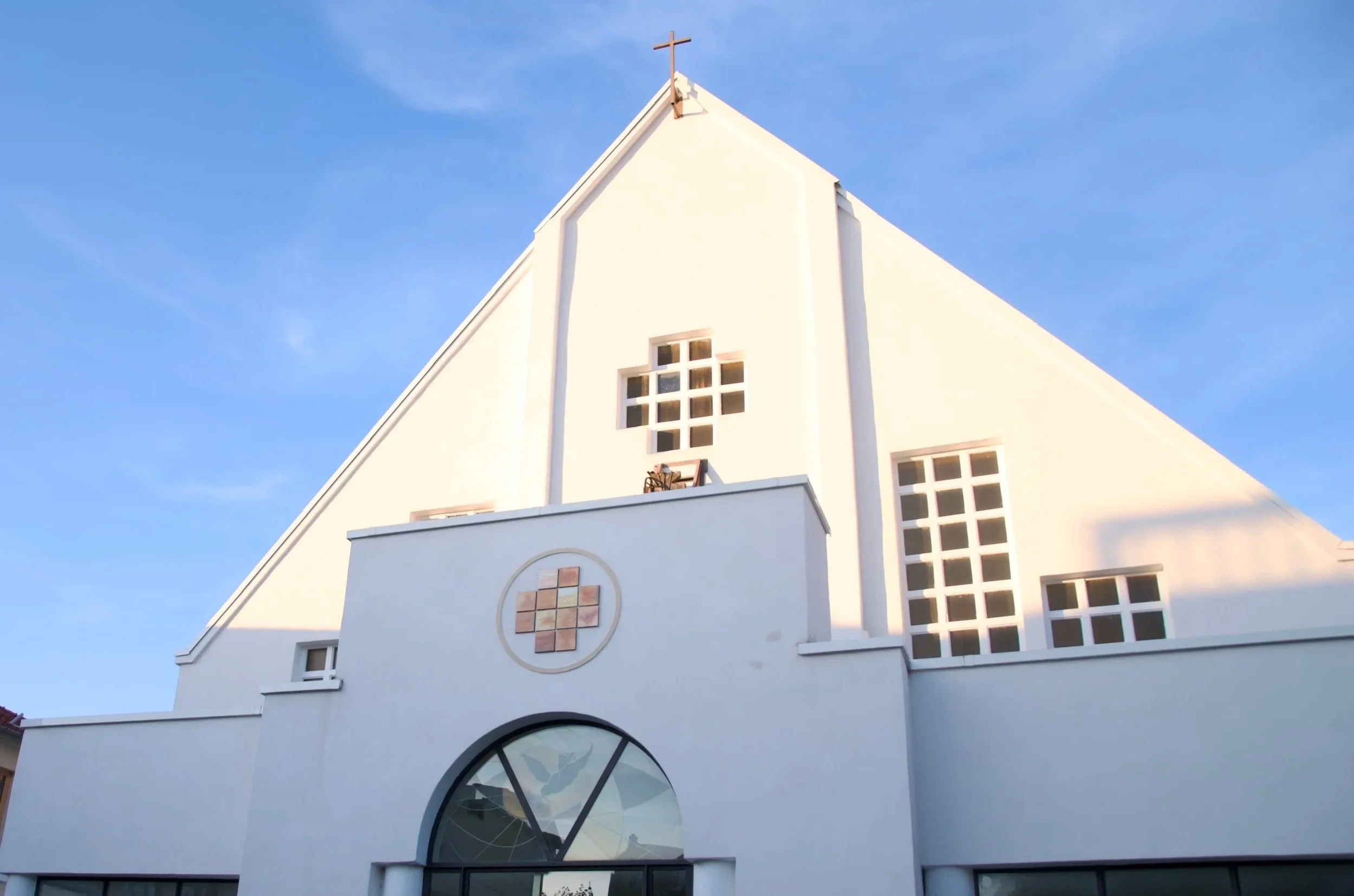 Une église blanche avec un toit en pente, une croix à son sommet, et des fenêtres à carreaux, sous un ciel bleu.