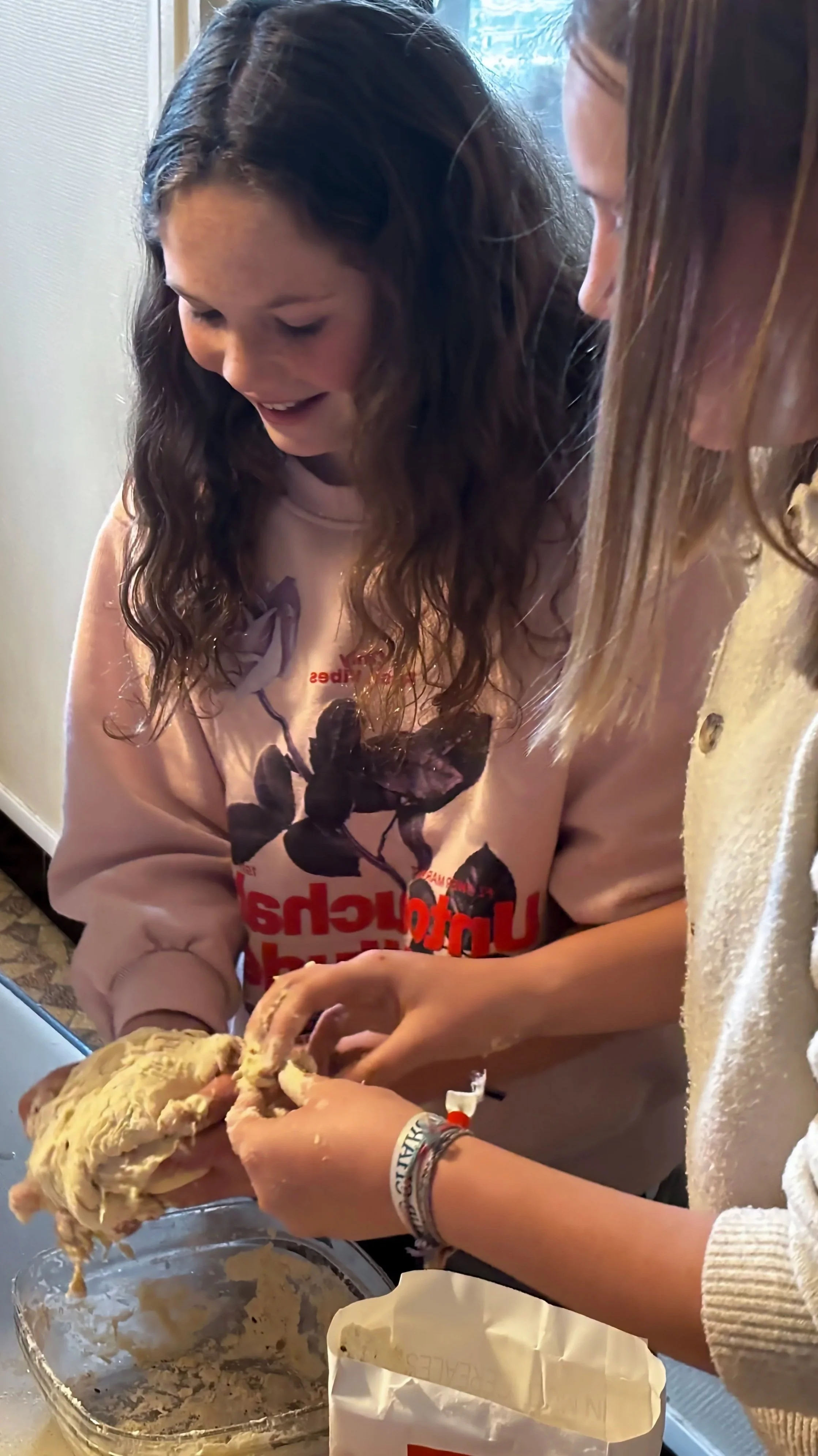 Deux jeunes filles préparent de la pâte dans une cuisine, avec des ingrédients et des ustensiles visibles sur la table.