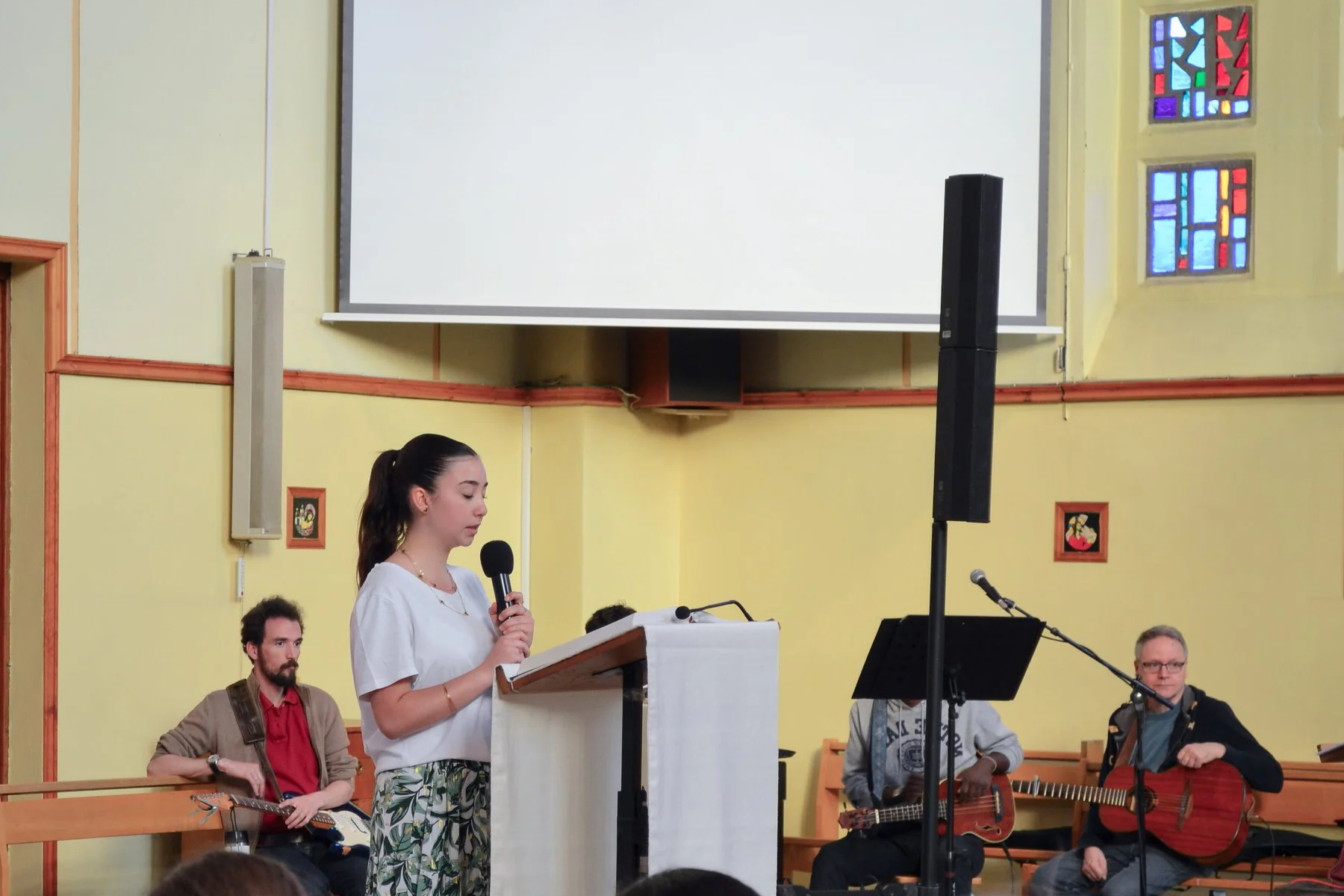 Une jeune femme parle dans un microphone lors d'une messe, entourée de plusieurs musiciens avec des guitares, dans une église avec de petites fenêtres colorées et des décorations murales. Un grand écran blanc est visible au fond.