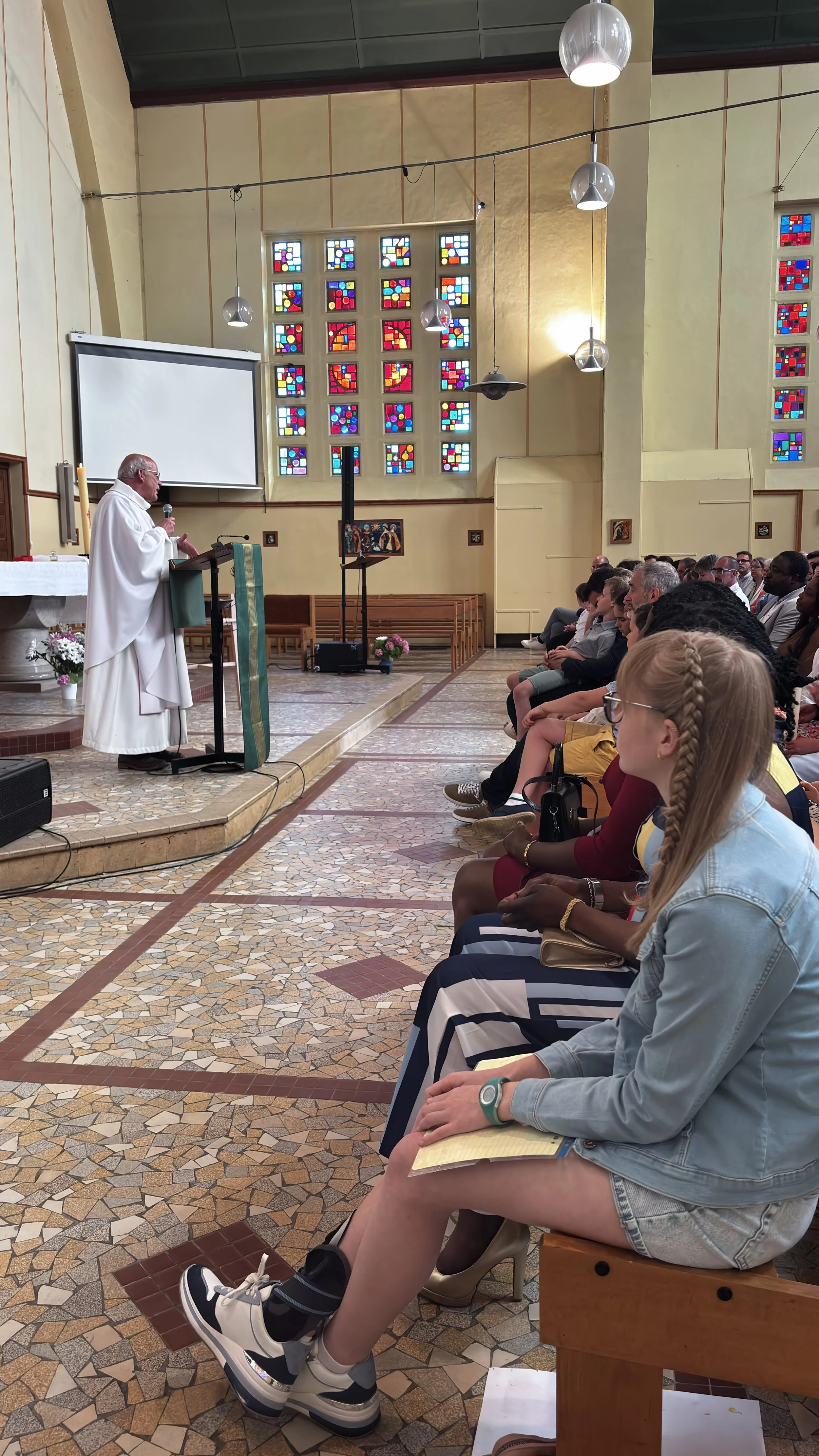Un prêtre en robe blanche parle à une assemblée assise dans une église avec des vitraux colorés en arrière-plan.