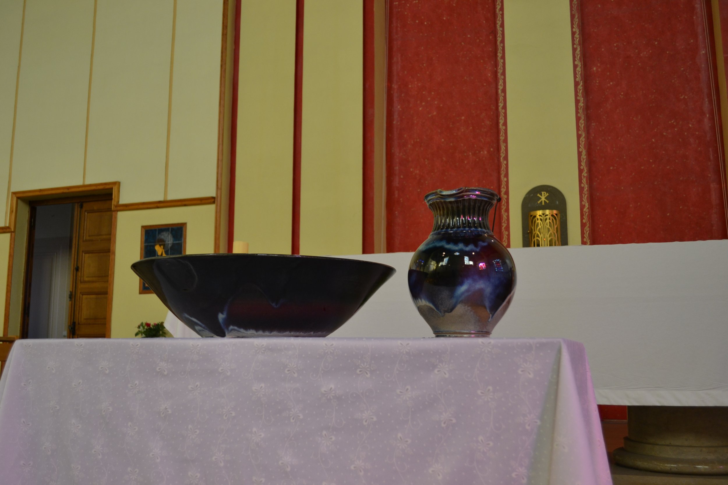 Coupes en céramique posées sur une table avec un drap blanc, dans une salle avec murs jaunes et rouges, décorée avec des œuvres , symbolisant le baptême chez les catholiques