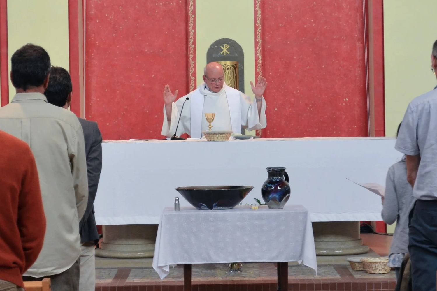 Un prêtre lors d'une messe catholique, levant les mains, derrière l'autel avec un calice et des livres, dans une église avec des plusieurs personnes debout en prière.
