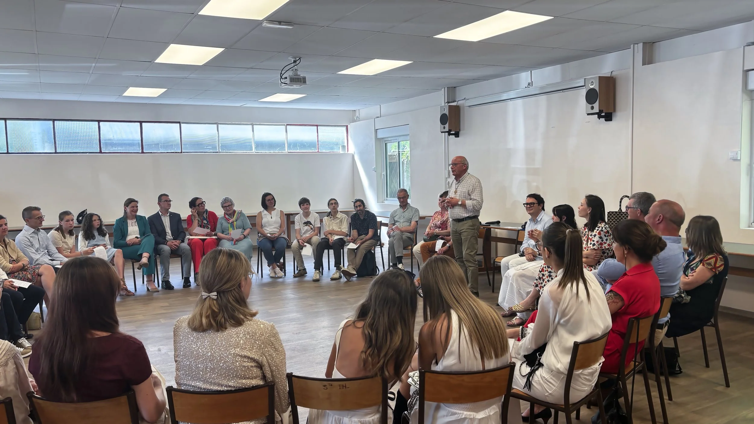 Une salle de conférence avec de nombreuses personnes assises en cercle, écoutant un homme qui parle debout, le prêtre fait donne une conférence. La pièce est bien éclairée avec de grandes fenêtres.