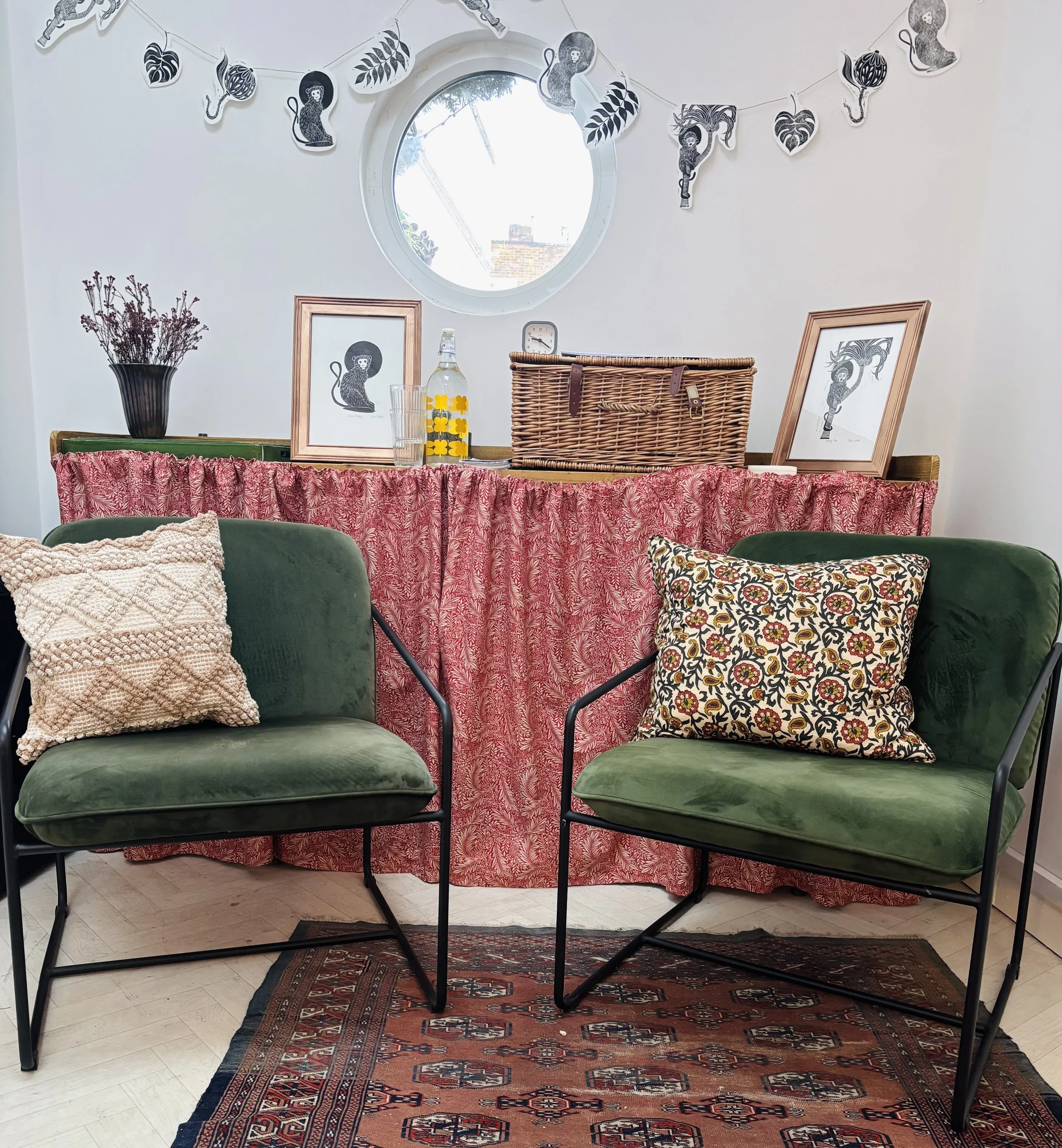 A cozy living space features two green velvet chairs with black metal frames, each adorned with a patterned throw pillow. Behind the chairs, a table with a red patterned skirt holds framed art prints, a wicker basket, a yellow and clear decorative bottle, and a vase with dried flowers. Above, a string of black-and-white animal and leaf banners decorates the white wall, with a round window providing natural light.