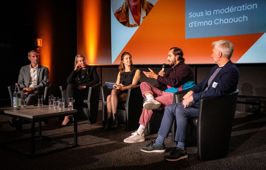 Panel discussion with five people seated on stage, one speaking into a microphone, during a conference with orange lighting and a presentation screen in the background.