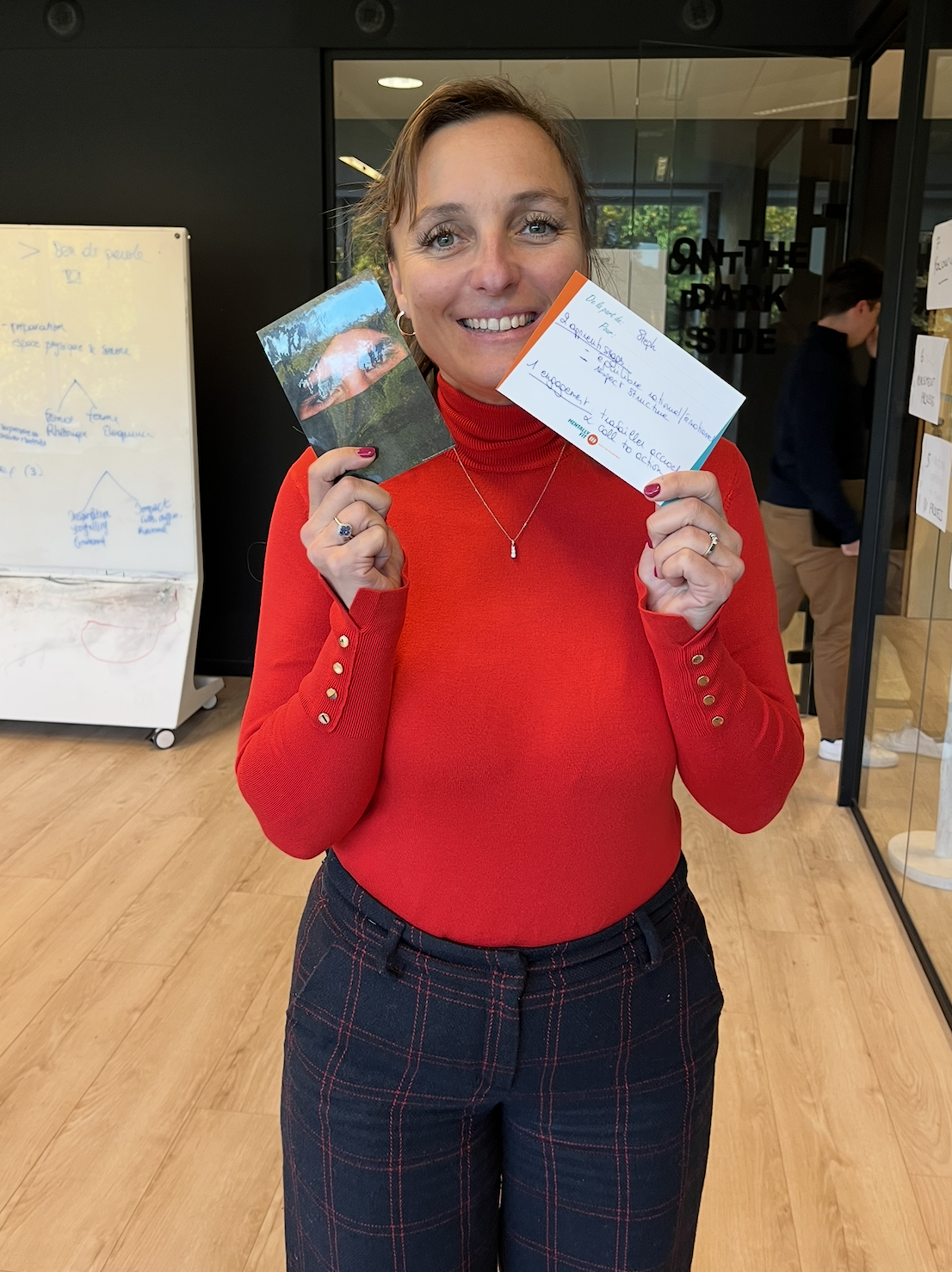 A person in a red sweater holding cards and smiling in an office setting with a whiteboard in the background.