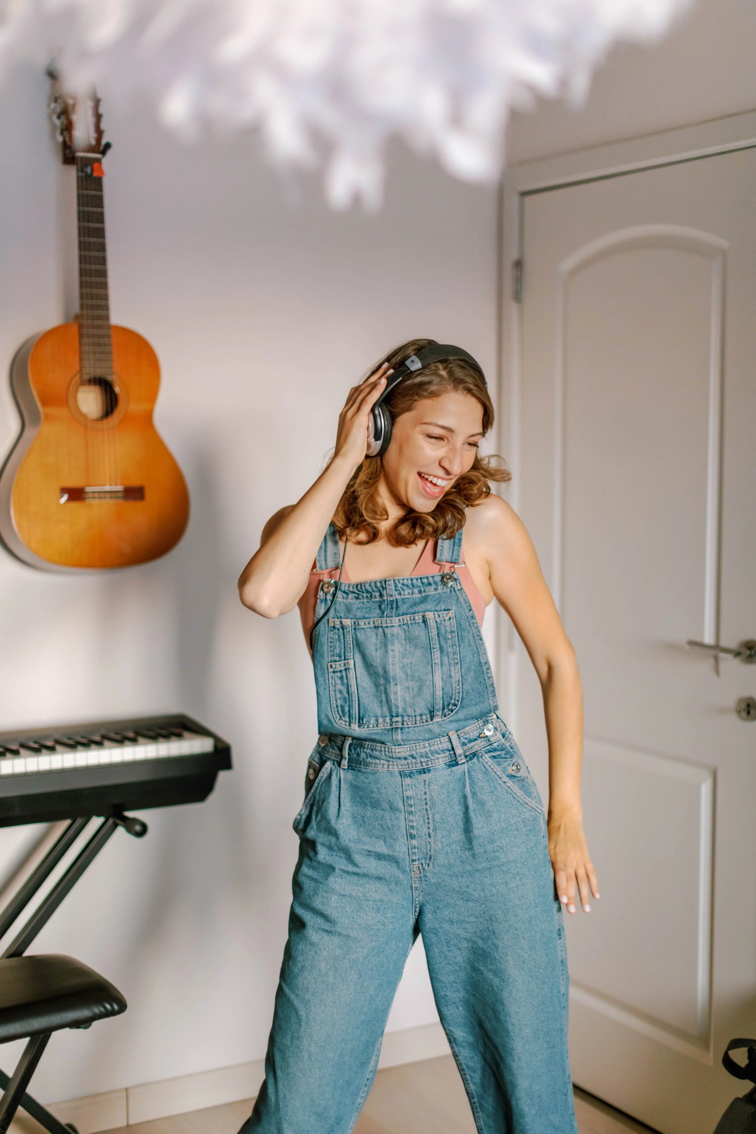 Woman in denim overalls listening to music with headphones, smiling and dancing in a room with a guitar on the wall and a keyboard nearby.