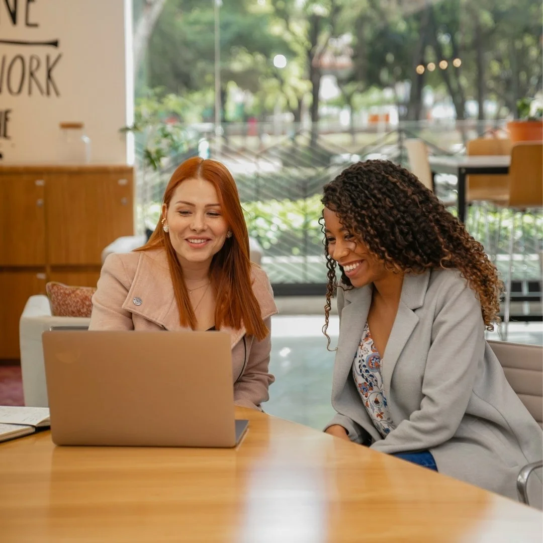 Two women smiling and looking at a laptop in an office setting.