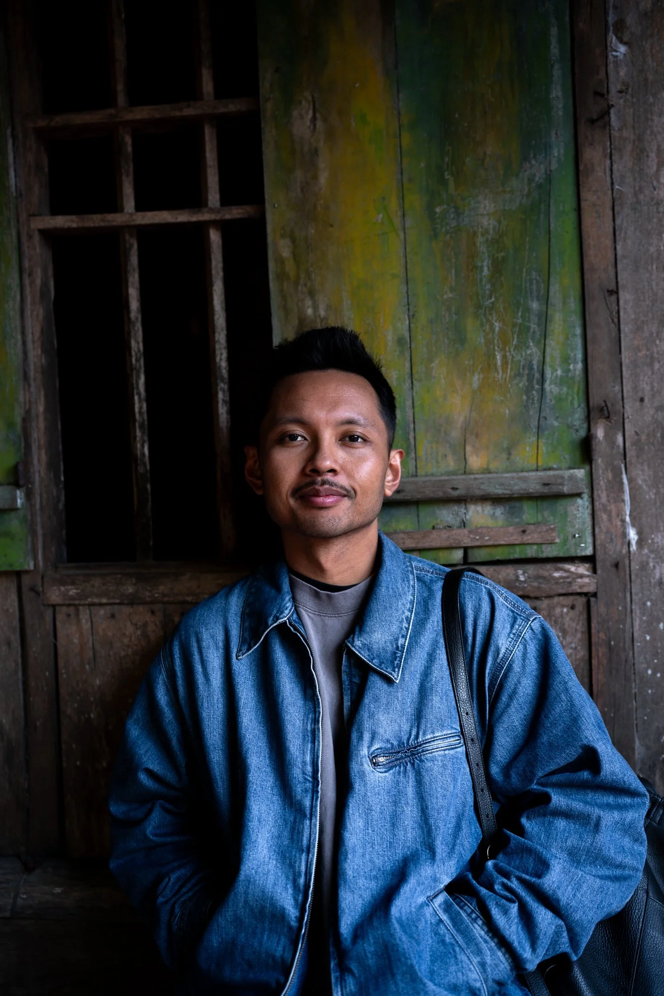 A young man with short black hair and a slight mustache, wearing a blue denim jacket over a gray shirt, stands in front of a rustic wooden wall with a small window and faded green paint.