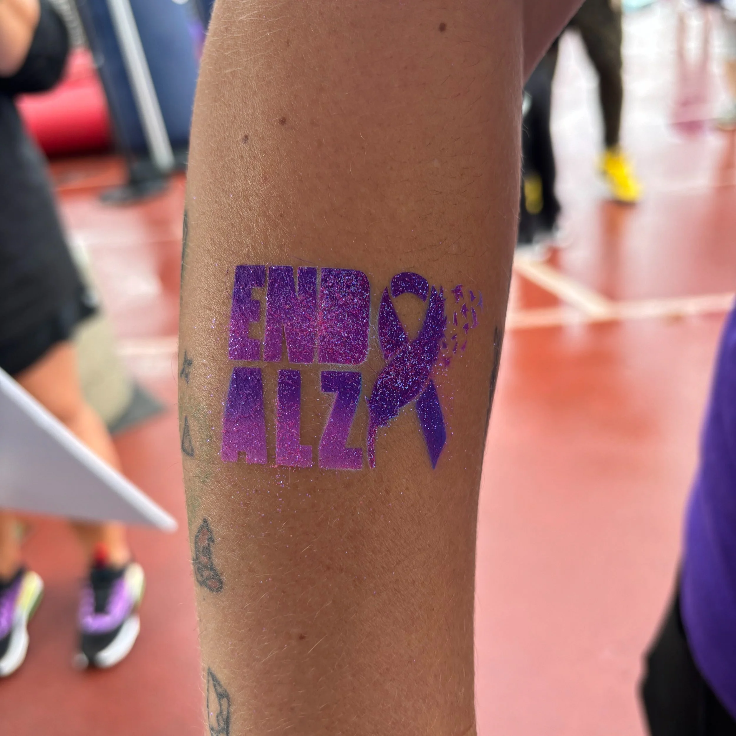 Purple glitter tattoo on arm with text 'END ALZ' and a ribbon, promoting Alzheimer's awareness, with blurred background of people. Bringing the excitement to events with our professional airbrush tattoo services—book today!