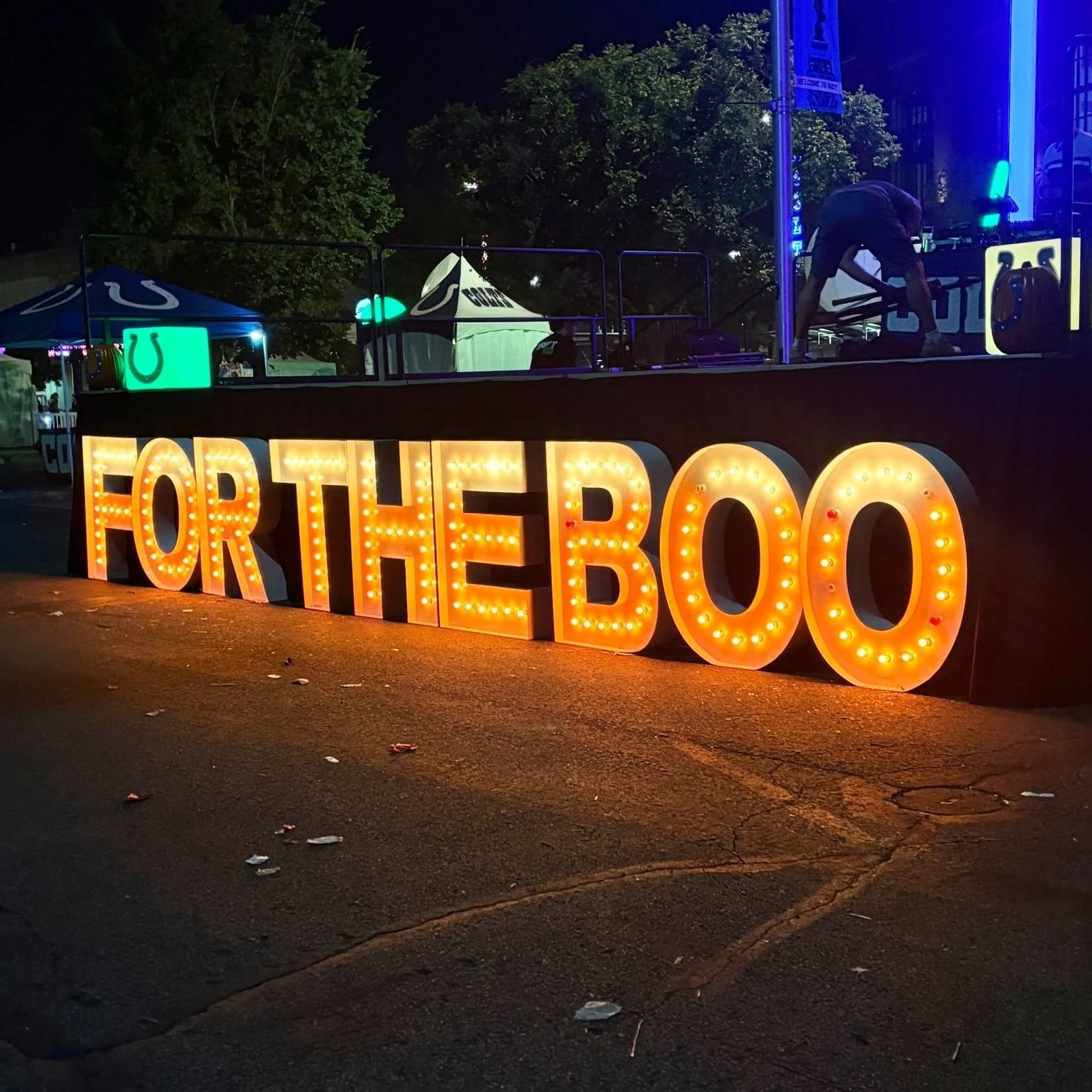 Illuminated sign with 'FOR THE BOO' at outdoor event, night setting with stage and tents in background. Bringing the excitement to events with our professional airbrush tattoo services—book today!