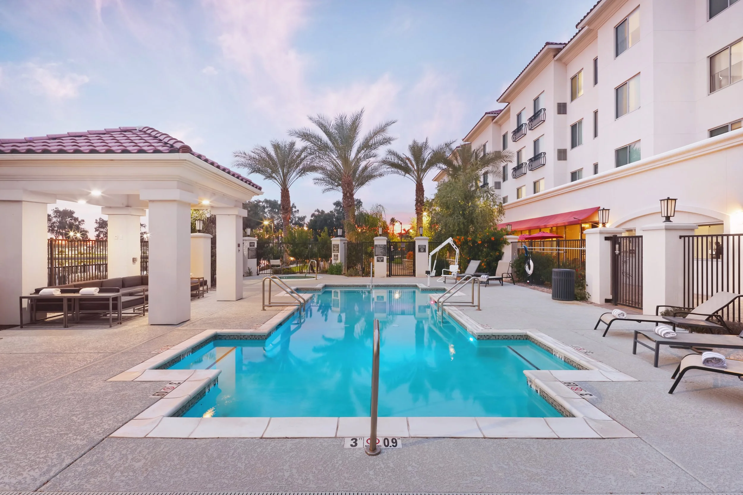 An outdoor swimming pool area at a hotel or apartment complex during early evening, with palm trees, lounge chairs, and a building with multiple stories in the background.