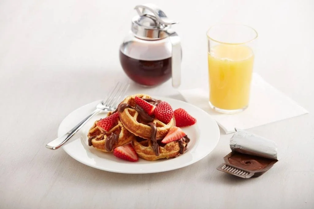 A breakfast plate with waffles topped with strawberries and chocolate drizzle, a glass of orange juice, a small jar of syrup, and a packet of chocolate spread on a white table.